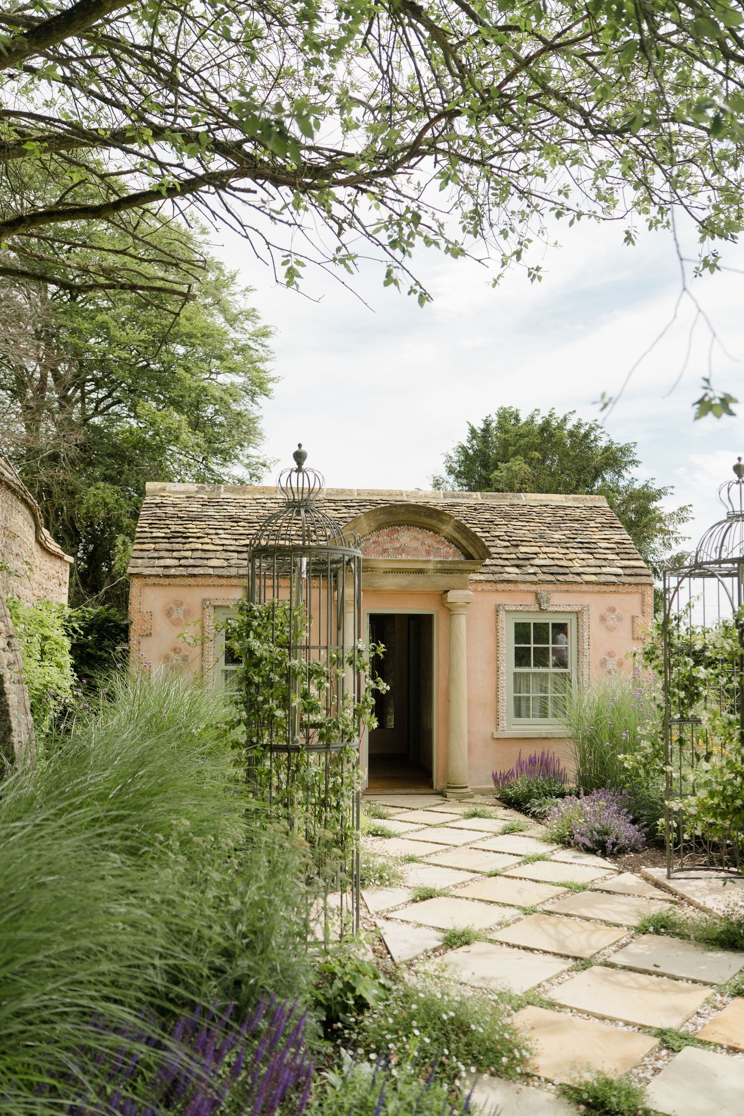 A charming pink stone cottage surrounded by lavender and lush greenery with a stone pathway leading to the entrance.