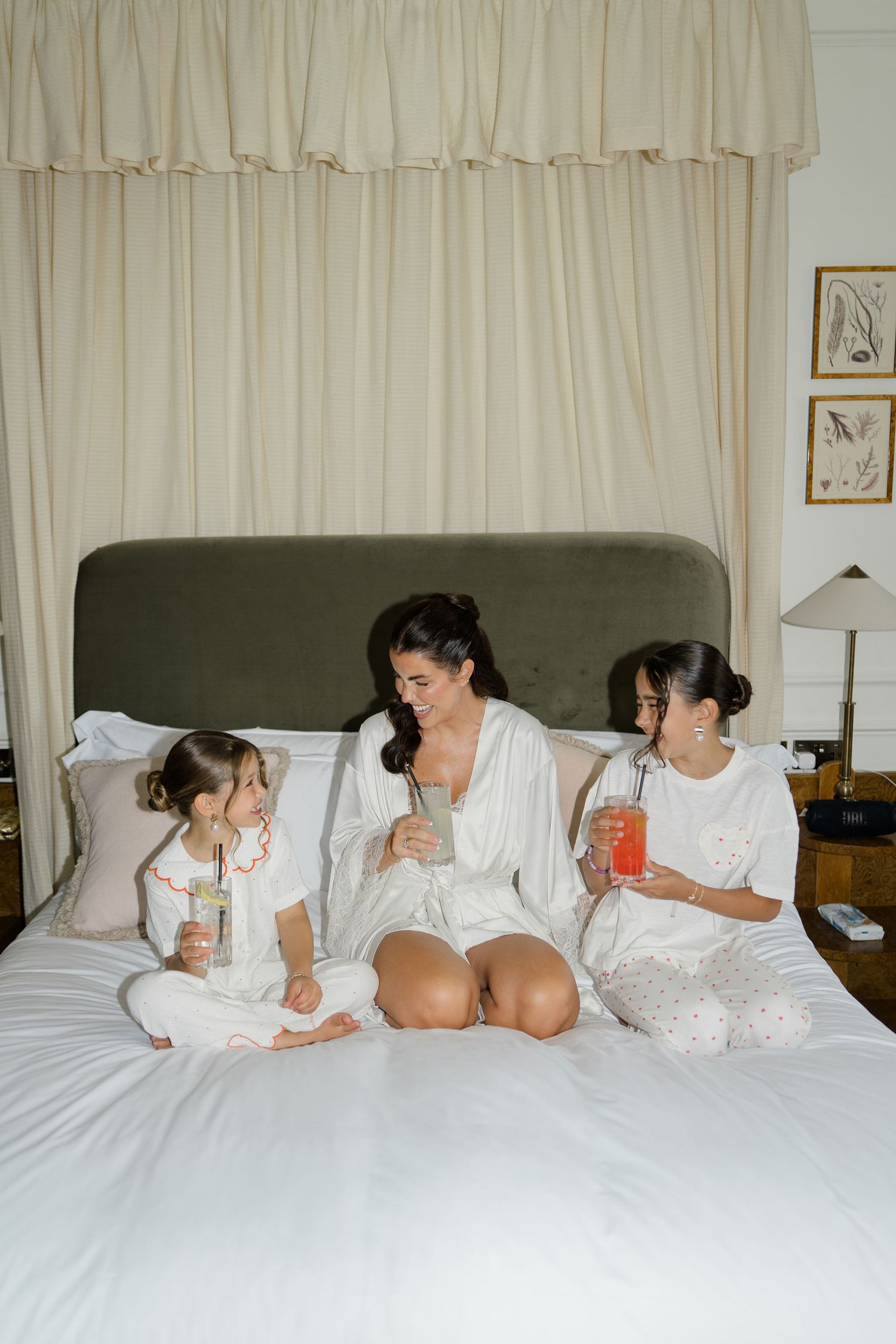 Bride in white robe sitting on bed with two young flower girls holding colorful drinks before wedding.