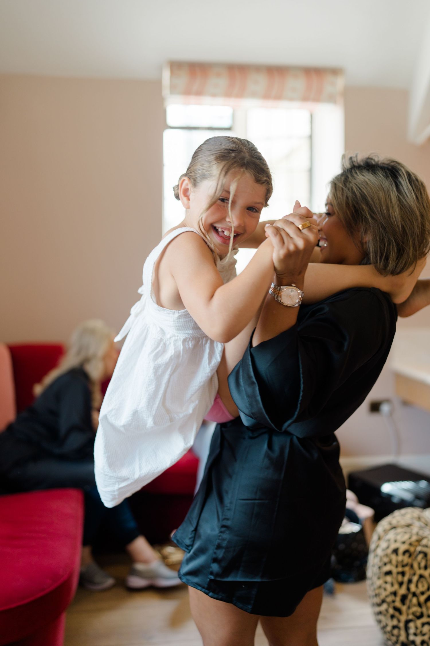 A joyful young flower girl in a white dress laughs while being held by a woman in a black satin robe.