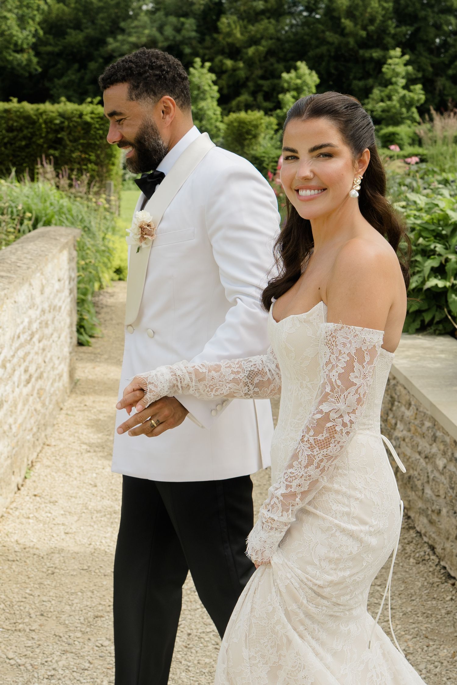 Bride in off-shoulder lace gown and groom in white tuxedo walking hand in hand in garden.