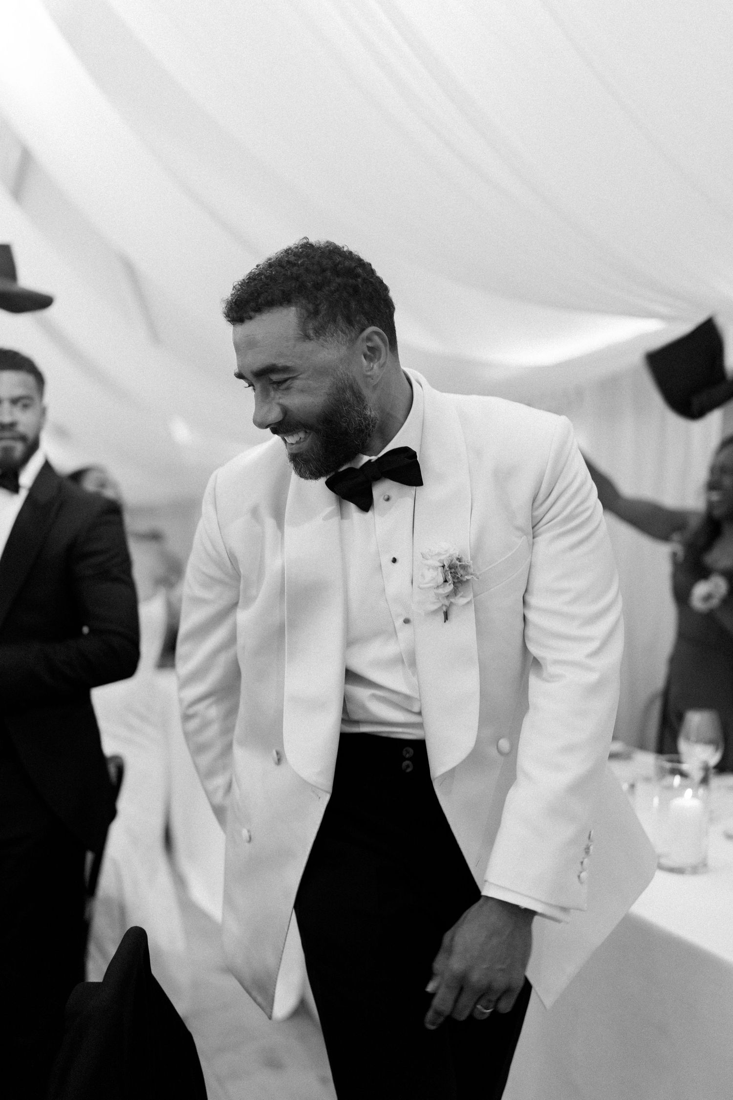Black and white photo of groom in white tuxedo jacket laughing joyfully inside wedding tent.