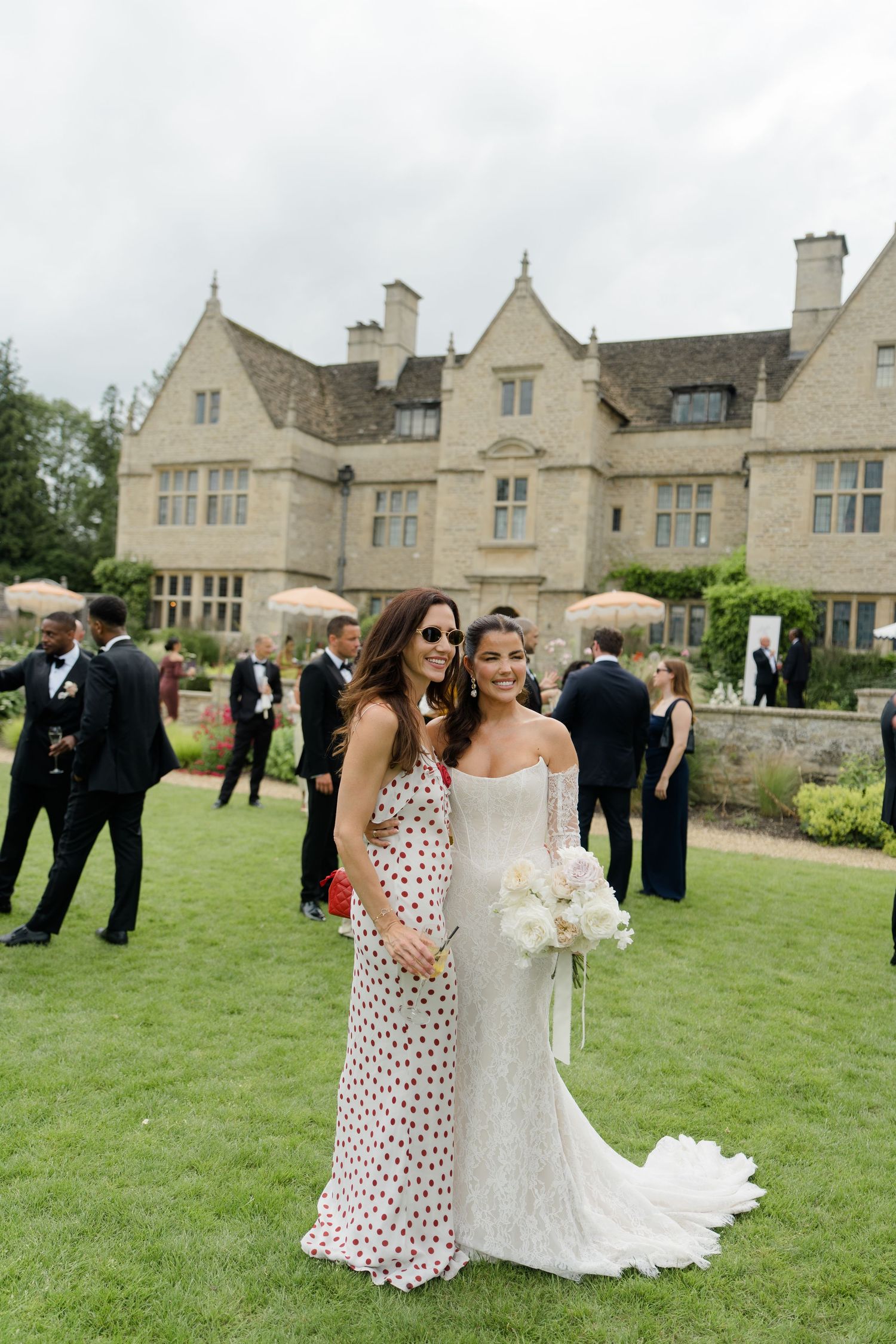 A bride in a lace gown holds white flowers while posing with a guest at a manor wedding.