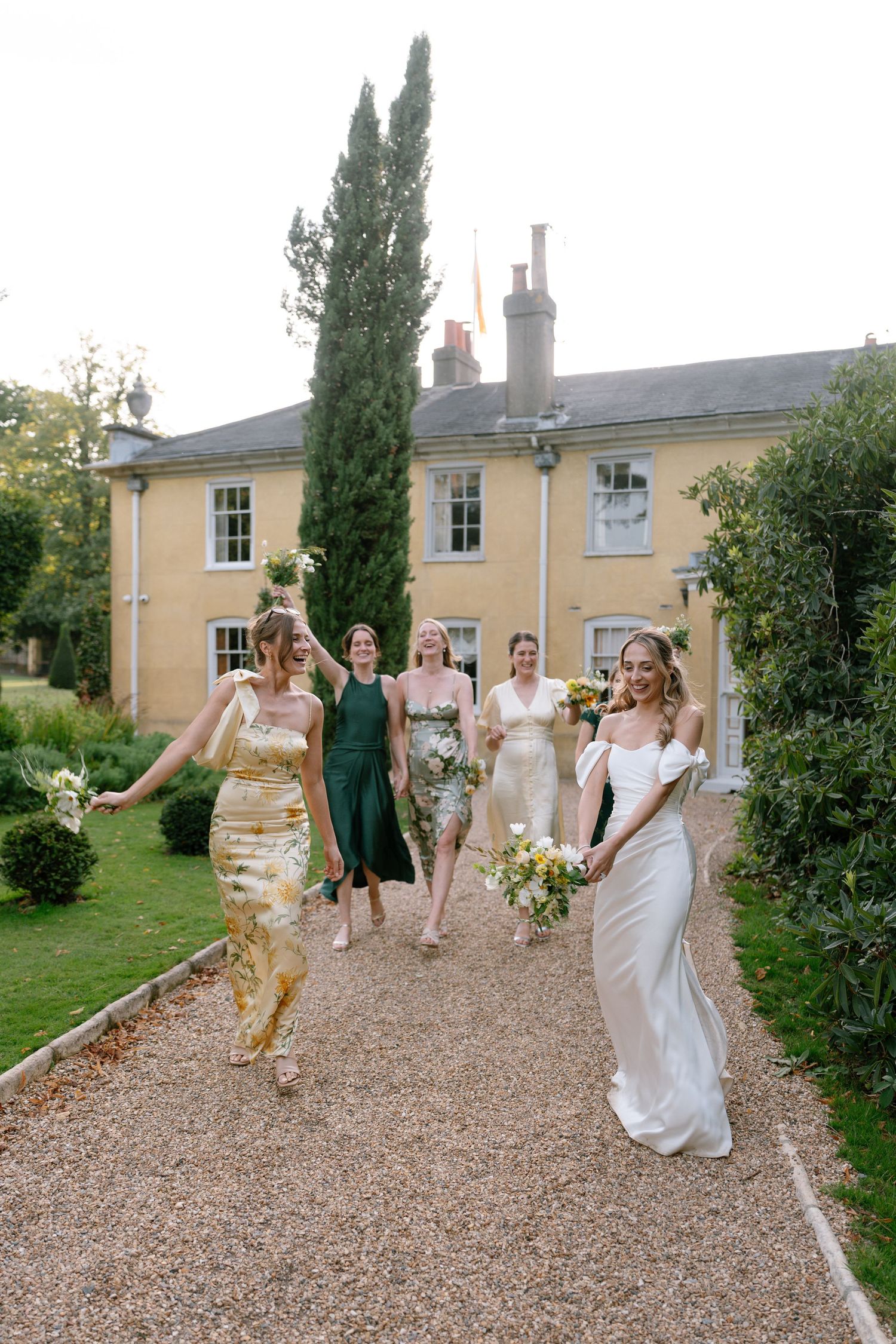 Wedding party walks and dances along a gravel path outside a yellow Georgian-style house.