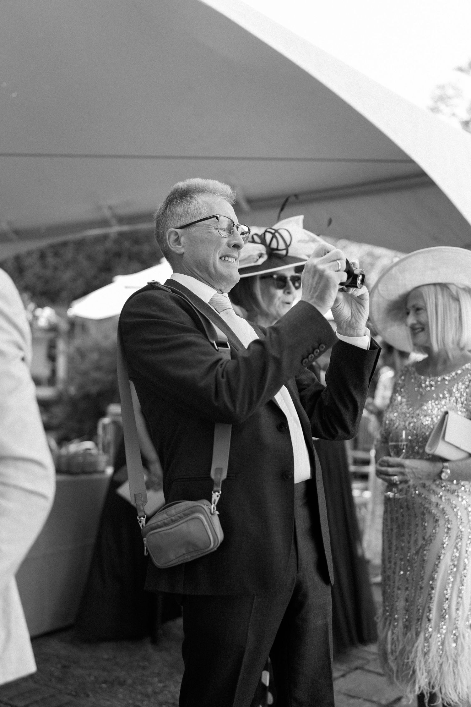Wedding guests in formal attire taking photos under a white tent.