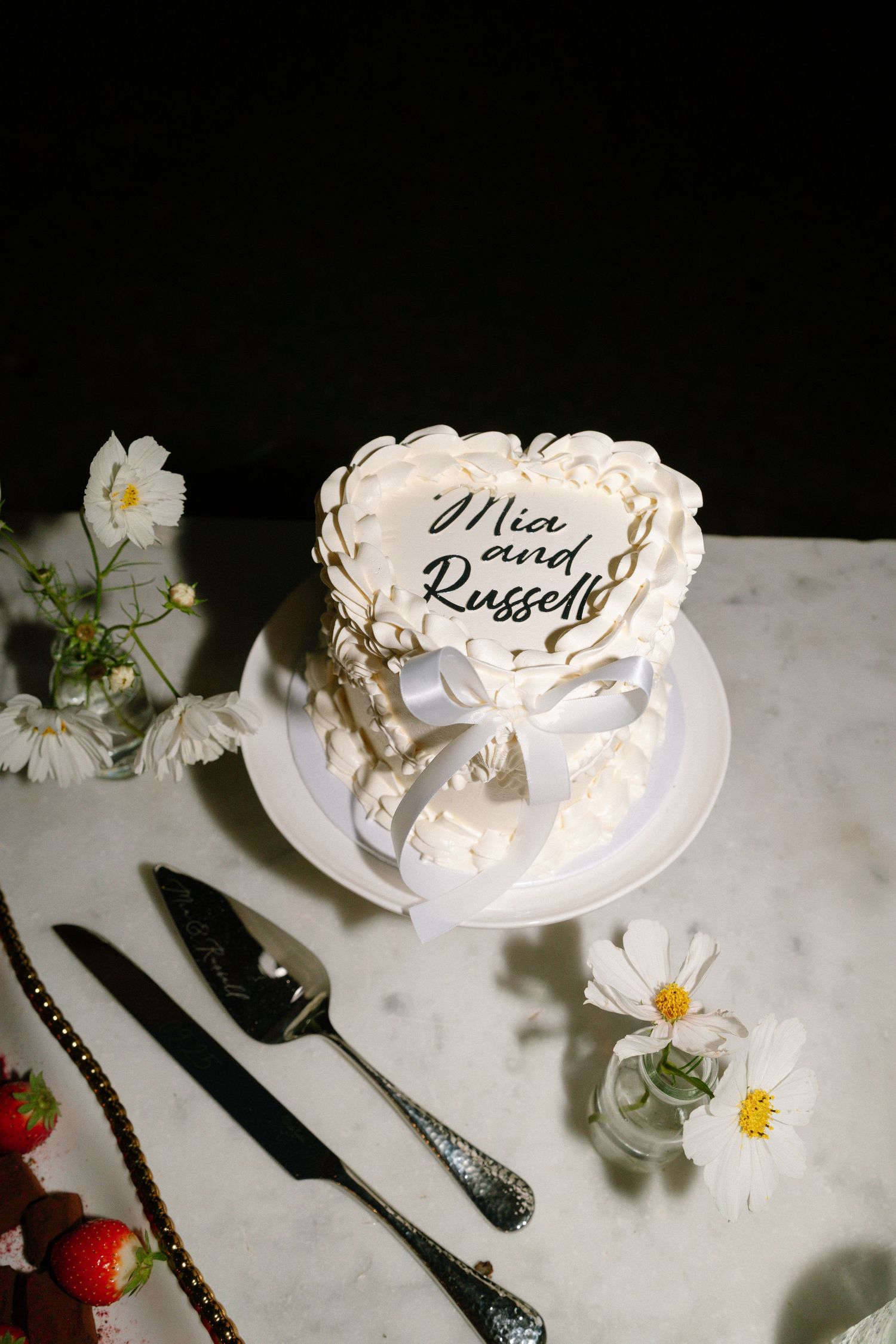 White wedding cake with decorative details and daisies on a table with silverware.