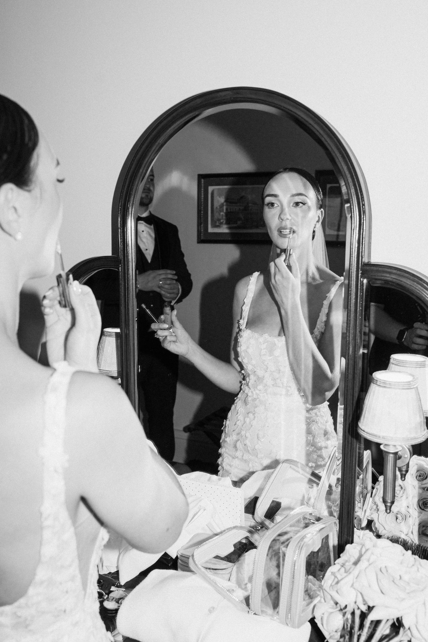 Bride getting ready in front of vanity mirror wearing white lace dress.