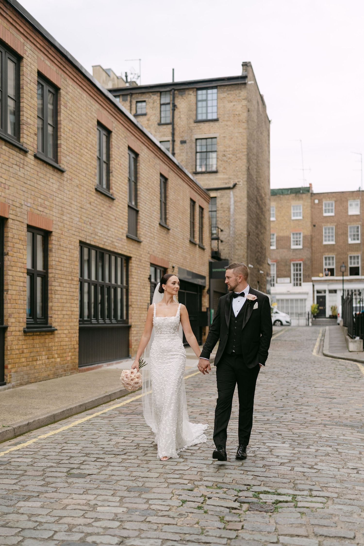 Newlyweds walk along a cobblestone street lined with brick buildings.