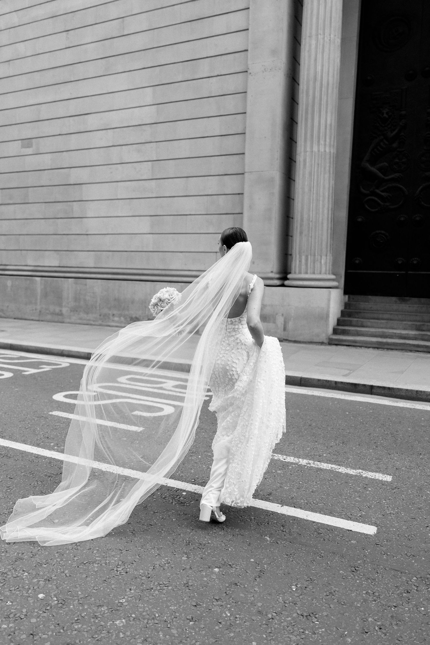 Black and white photo of bride's veil flowing dramatically on urban street.