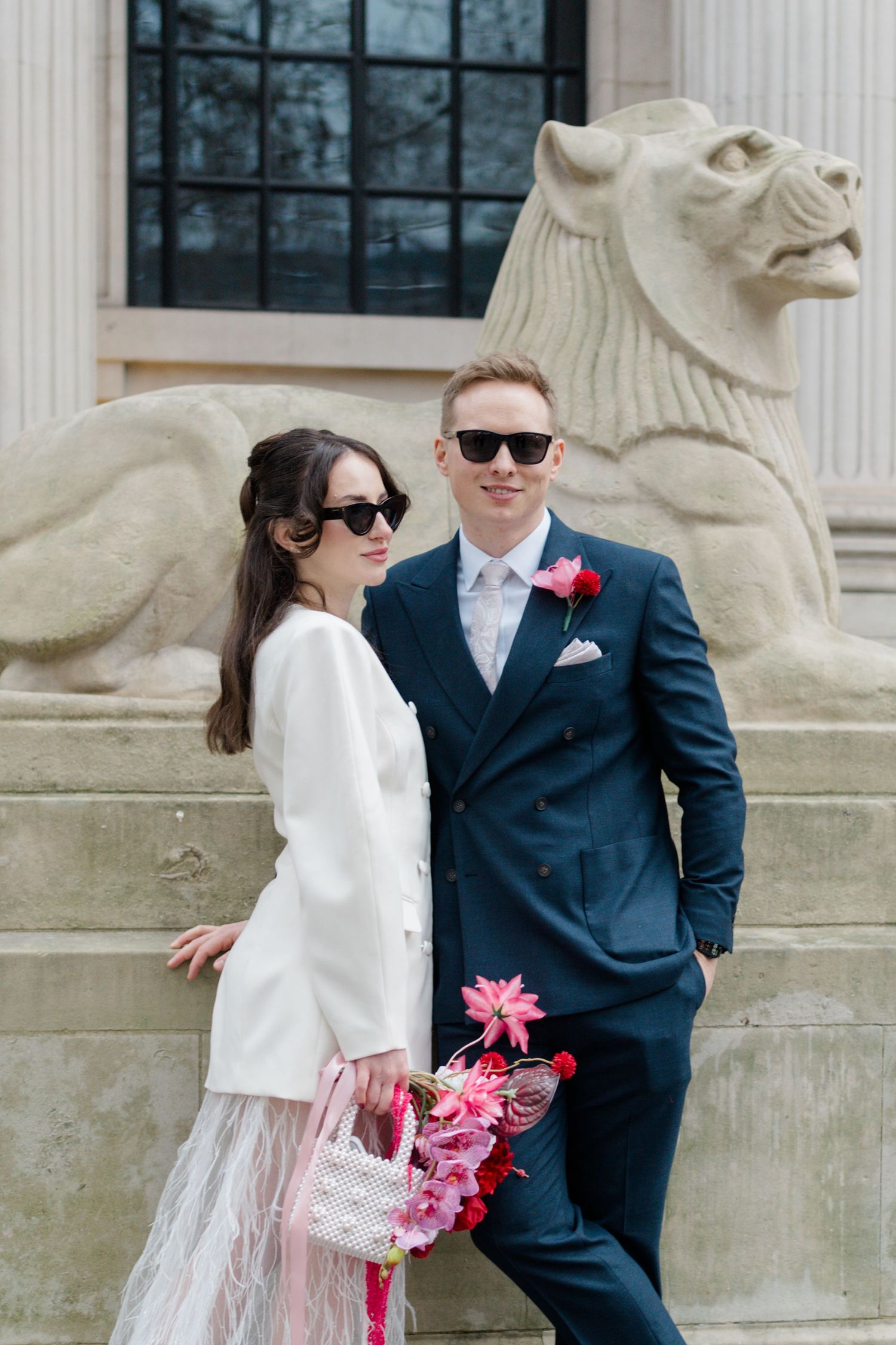 Stylish couple in sunglasses posing before stone lion sculptures, bride in white jacket with pink bouquet, groom in navy suit.