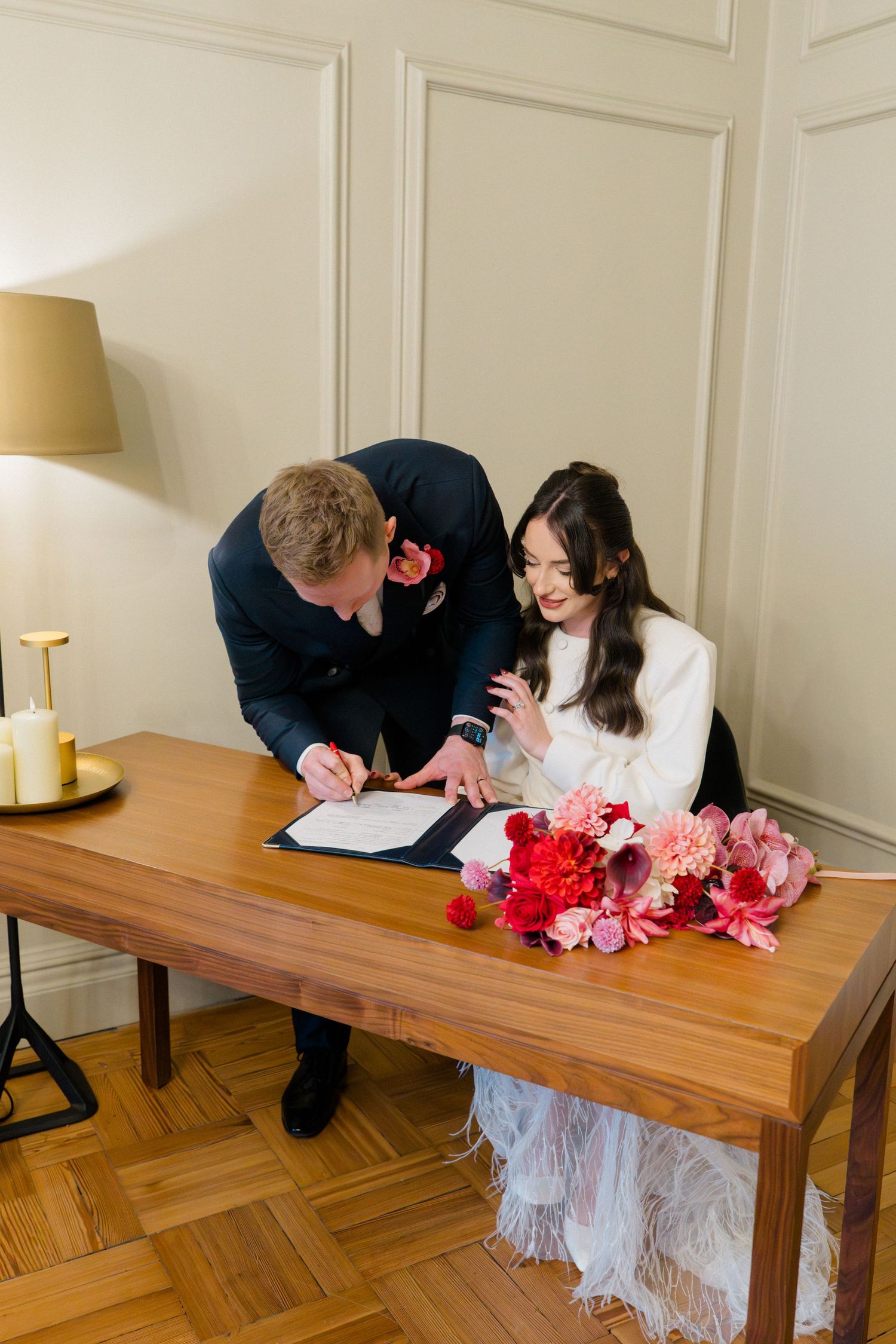 Couple signing wedding register at wooden table adorned with vibrant pink and red floral bouquet.