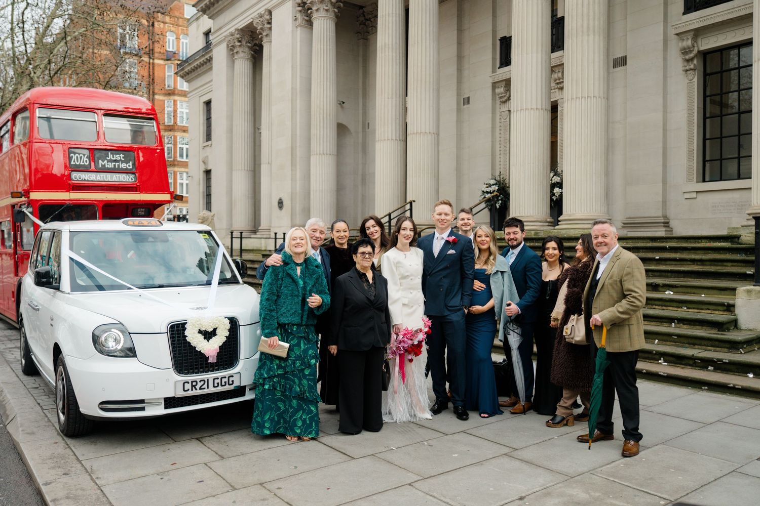 Wedding party poses outside a London registry office with a white taxi and red double-decker bus.