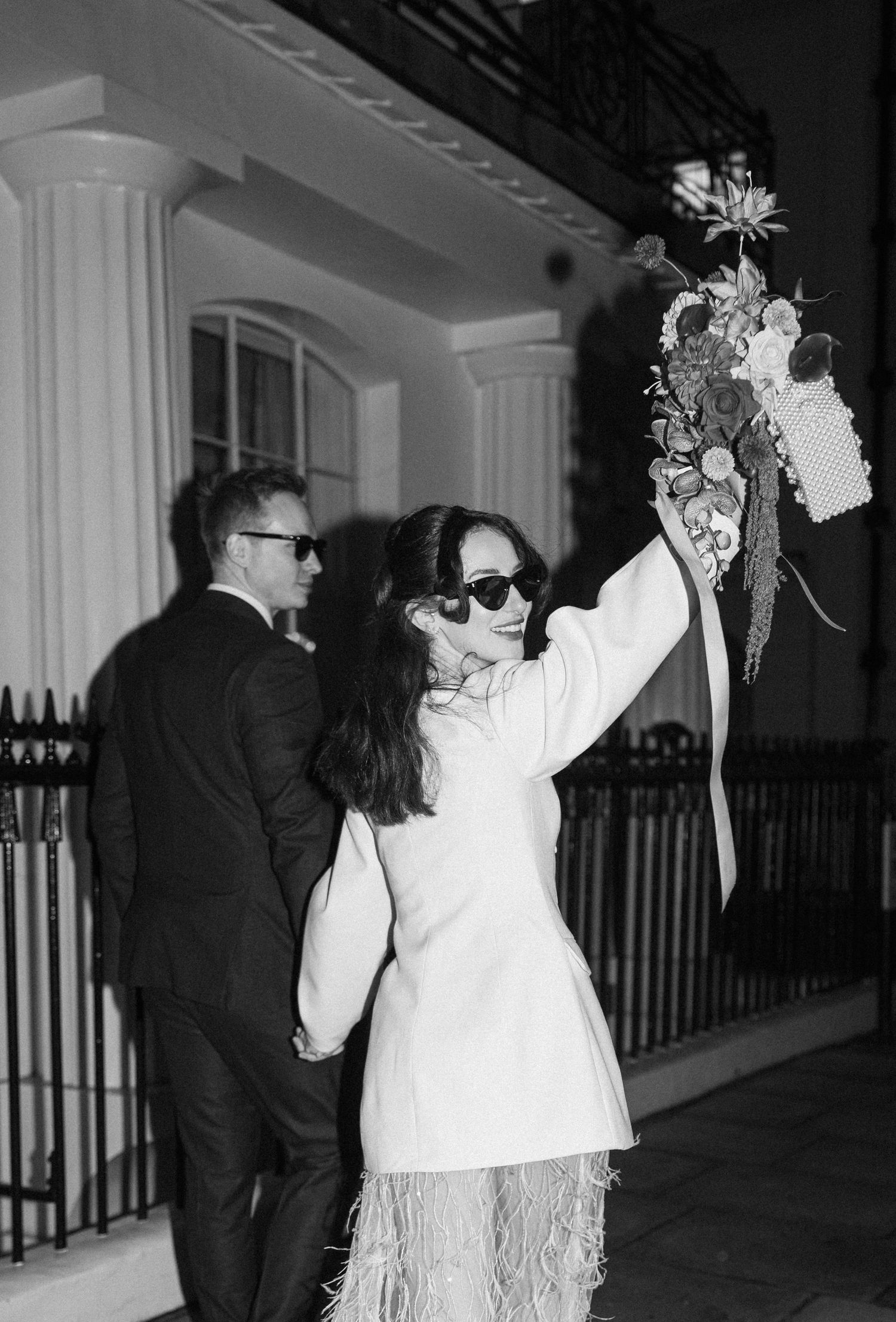 Black and white photo of bride in white feathered mini dress raising bouquet outside at night.