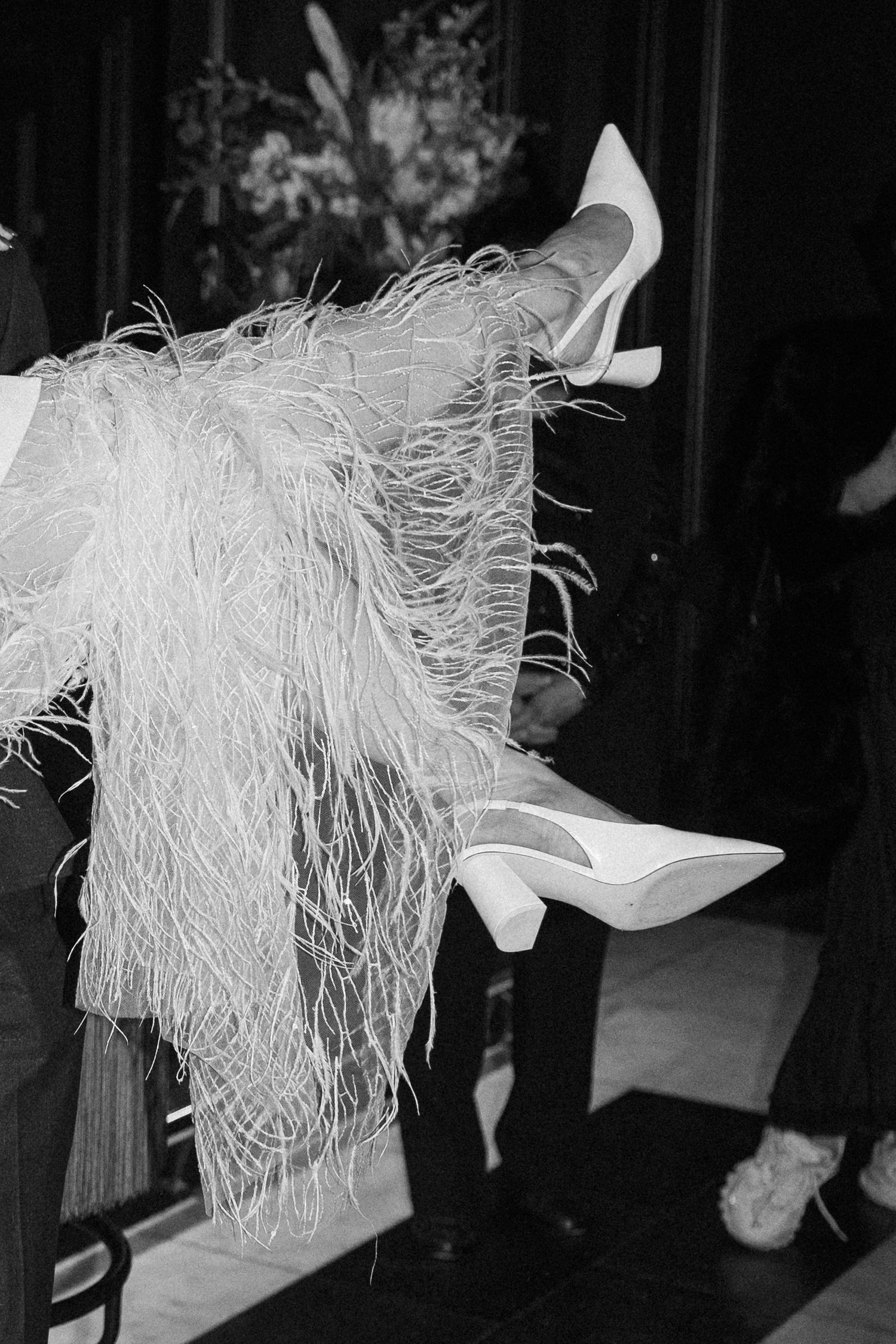 Black and white close-up of bride's white feather skirt hem and pointed white heels at wedding venue.