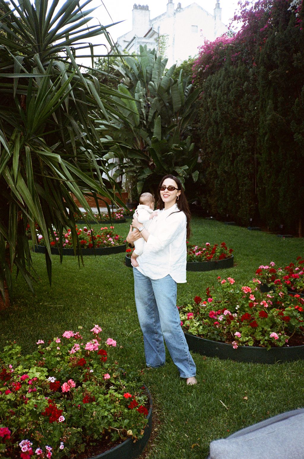 Woman in white shirt and jeans holds baby in lush garden with colorful flower beds and tropical plants.