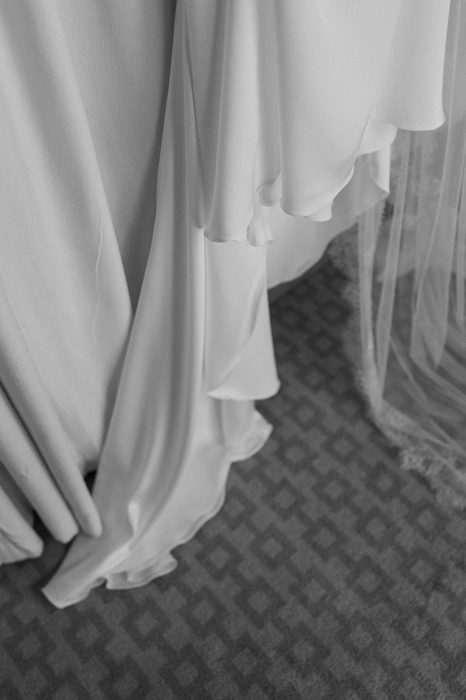 Black and white close-up of a flowing white wedding dress and tulle veil pooling gracefully on a patterned carpet.