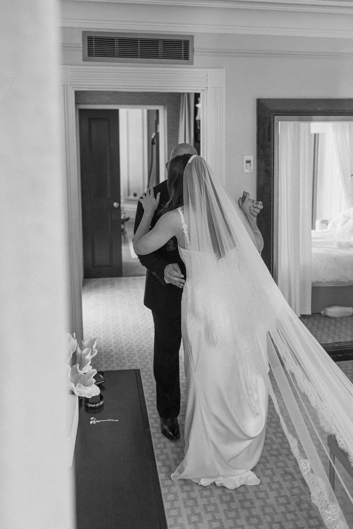 Black and white photo of bride in long veil embracing groom from behind in elegant hotel room.
