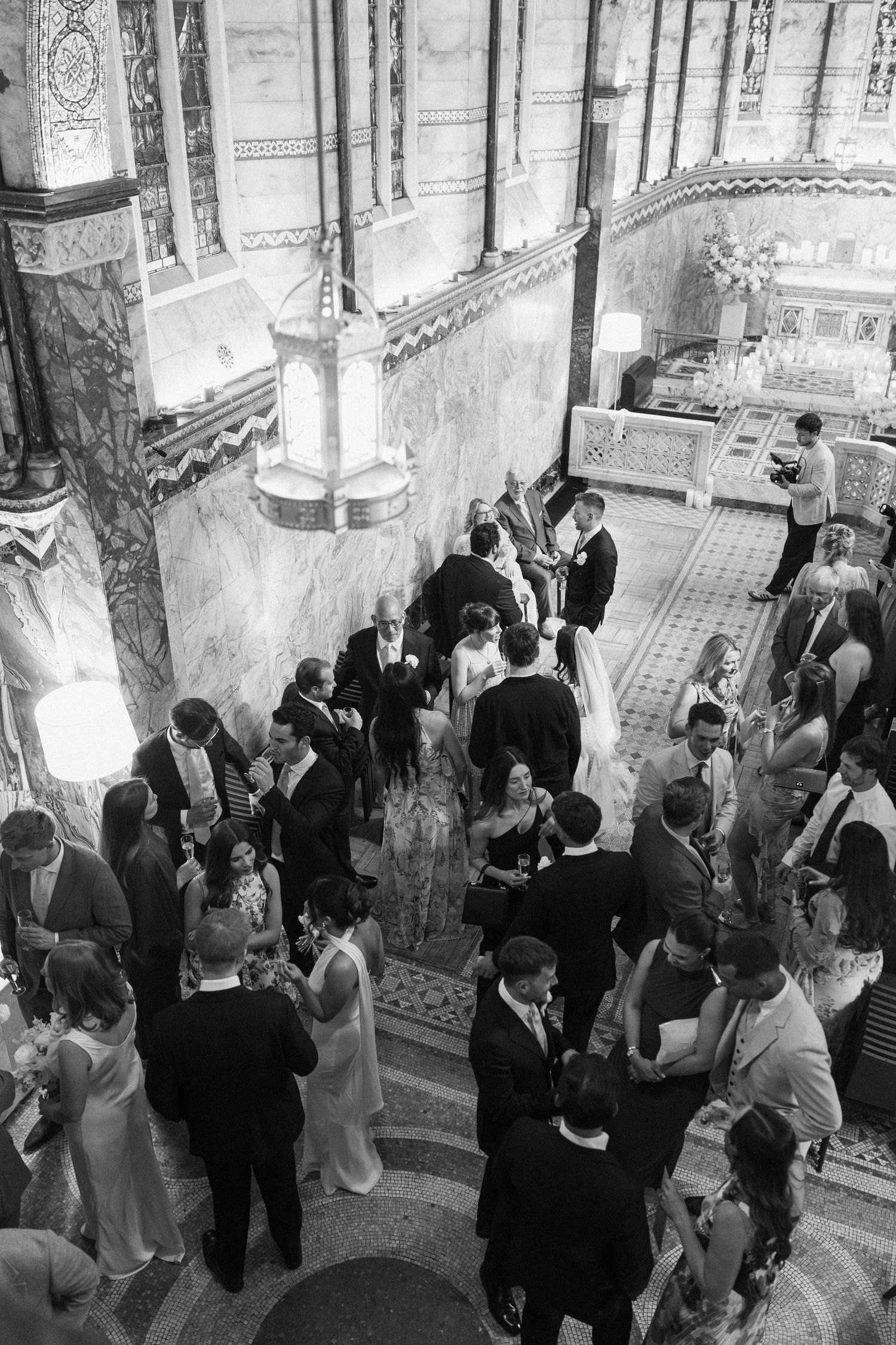 Aerial black and white view of wedding guests mingling in ornate marble church interior with chandeliers.