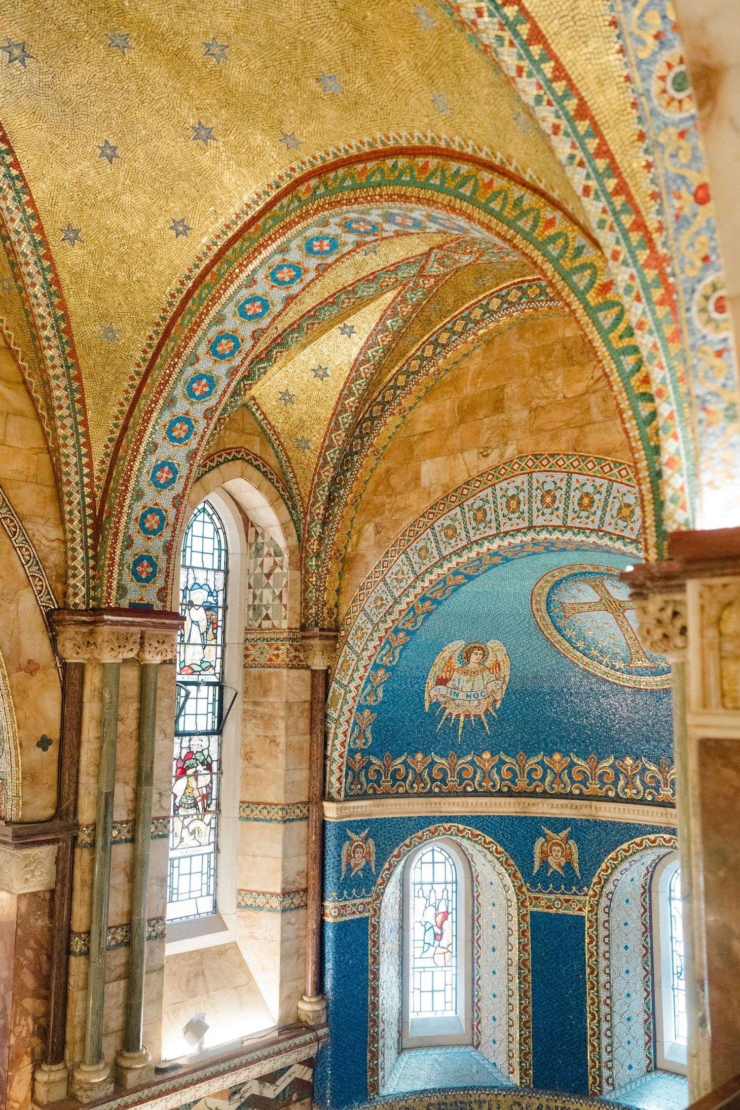 Ornate church interior featuring golden mosaic ceiling, blue apse artwork, and colorful stained glass windows.
