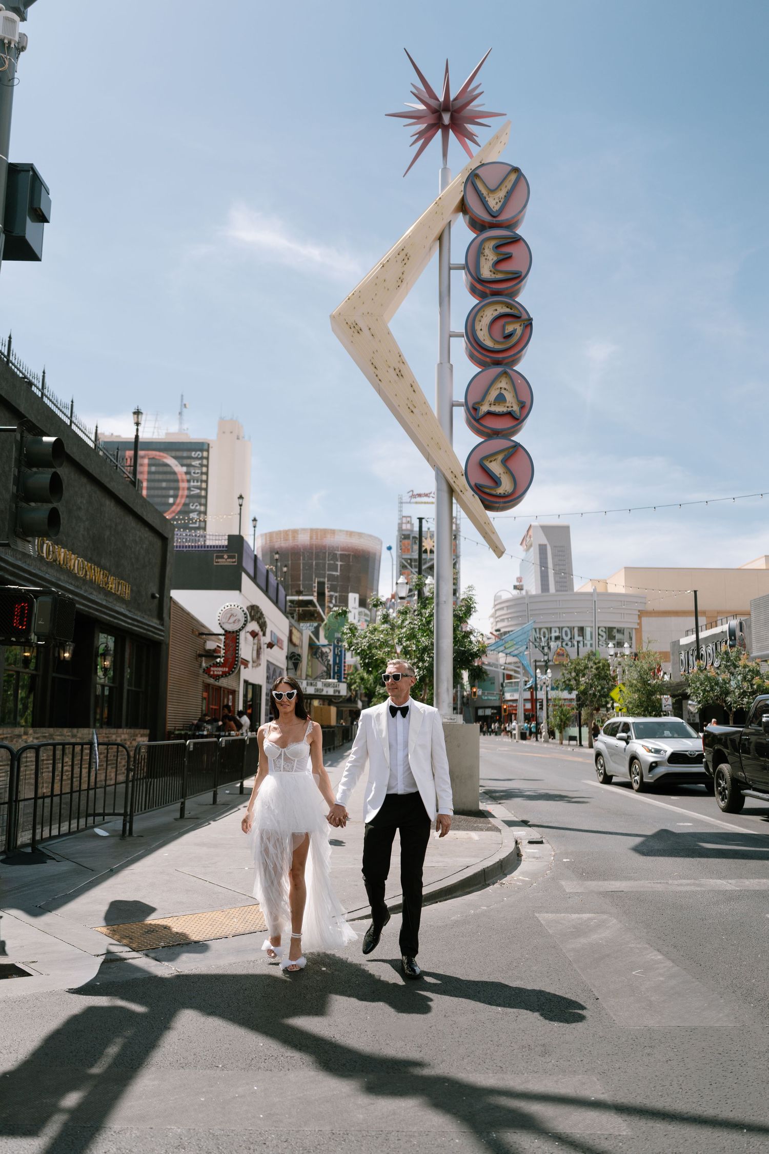 Couple holding hands while walking under vintage Vegas sign on sunny day.