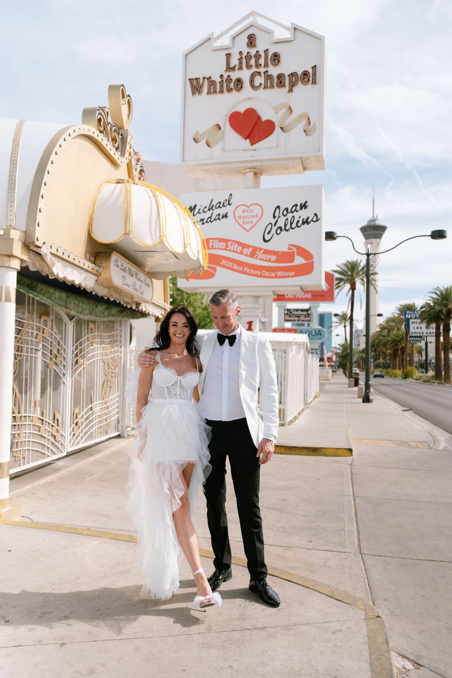 Happy couple poses for photos outside the Little White Chapel with palm trees in background.