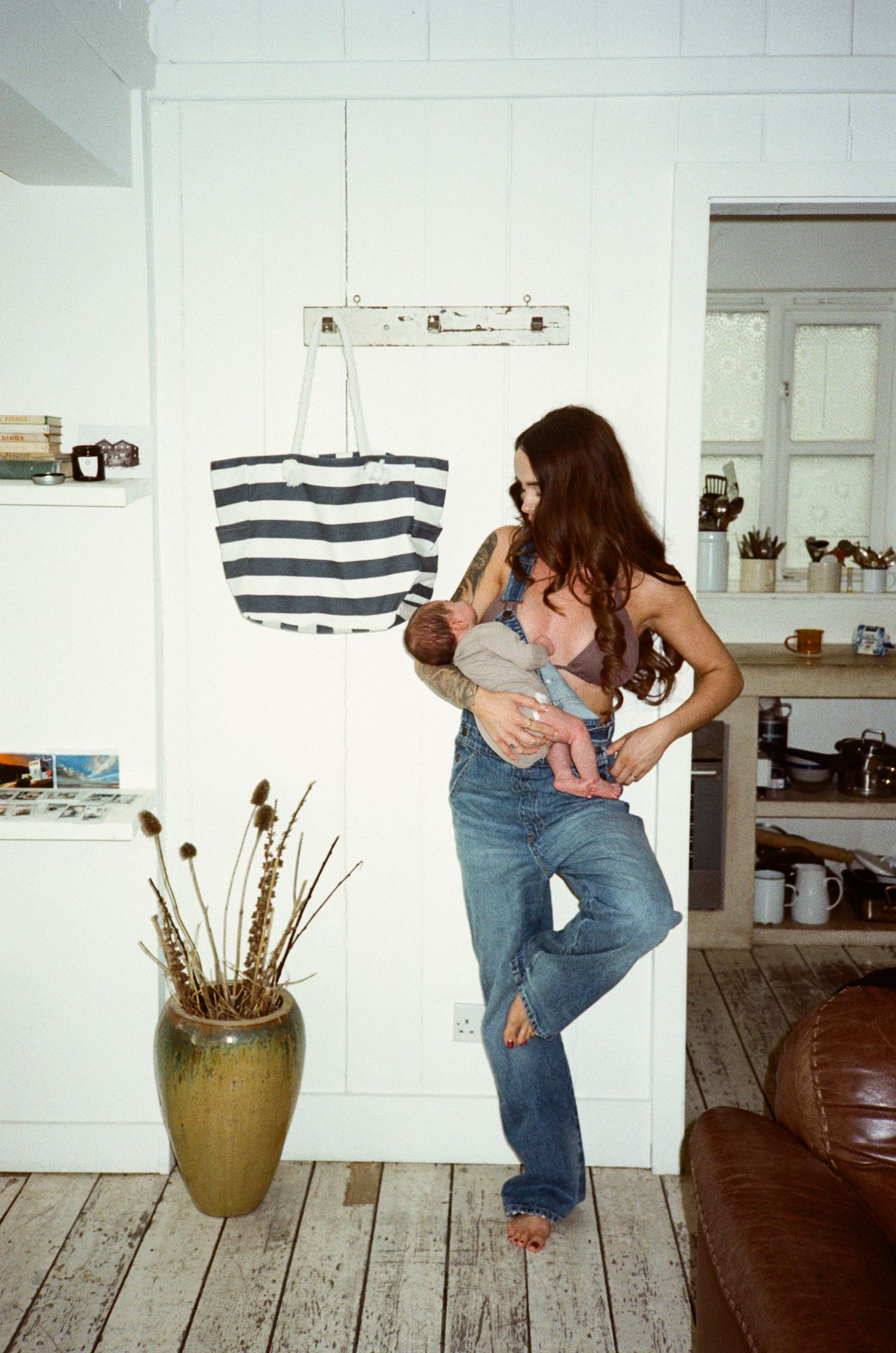 Young tattooed mother in denim overalls holding newborn baby in bright white bohemian home interior.