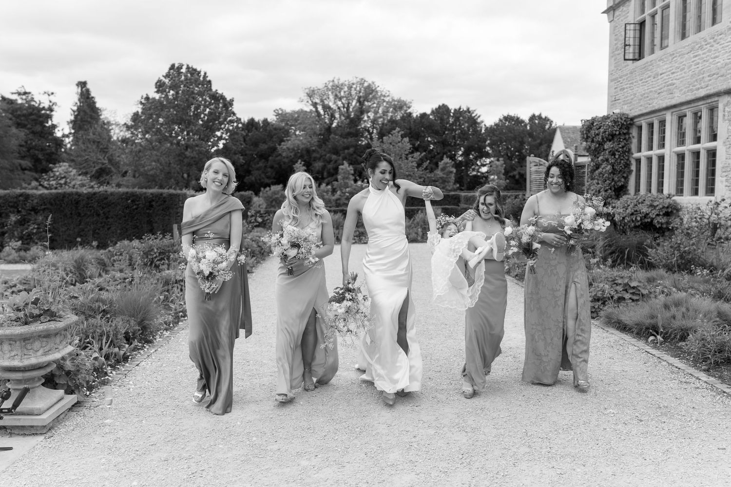 Bride and four bridesmaids in matching grey gowns walking through garden in black and white photo.