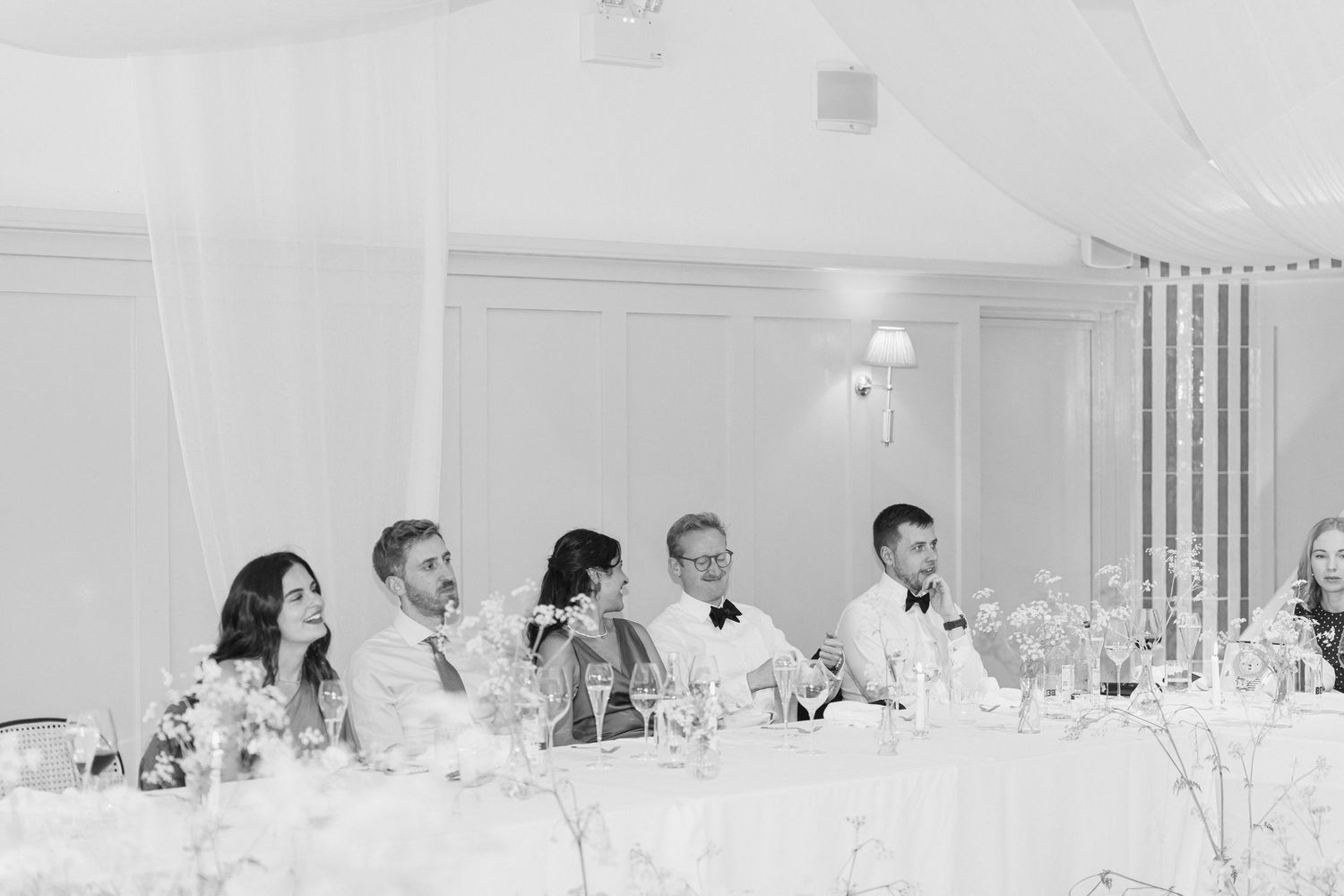 Wedding party seated at head table during reception in elegant white draped venue, black and white.