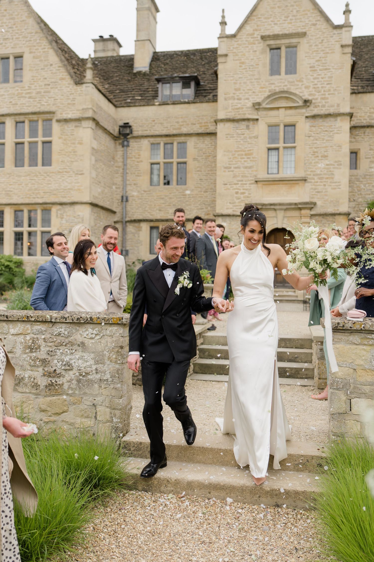 Bride and groom descend stone steps at a Cotswolds manor house wedding, greeted by confetti from smiling guests.