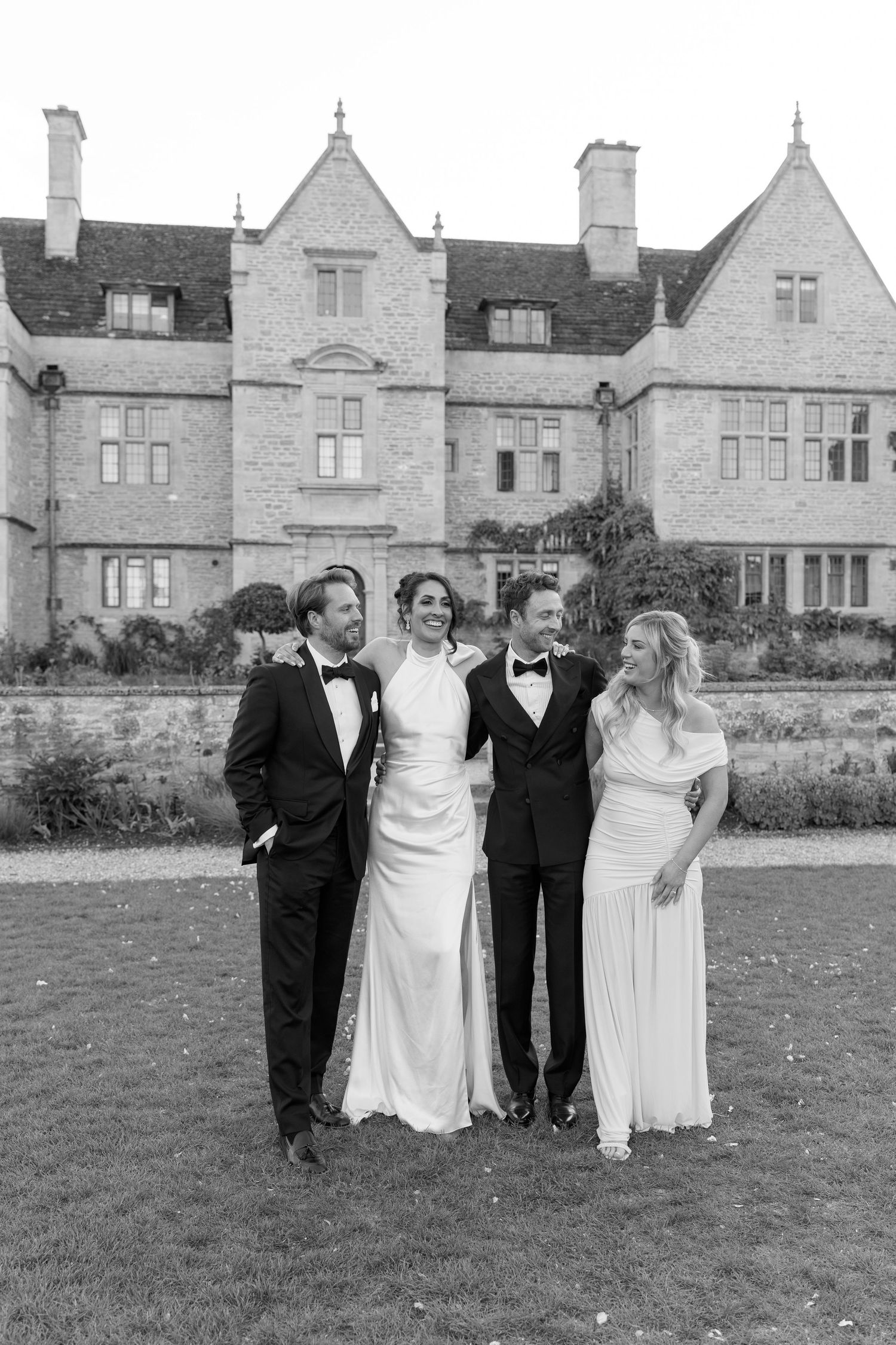 Black and white portrait of bride, groom and two guests posing in front of grand stone manor house.