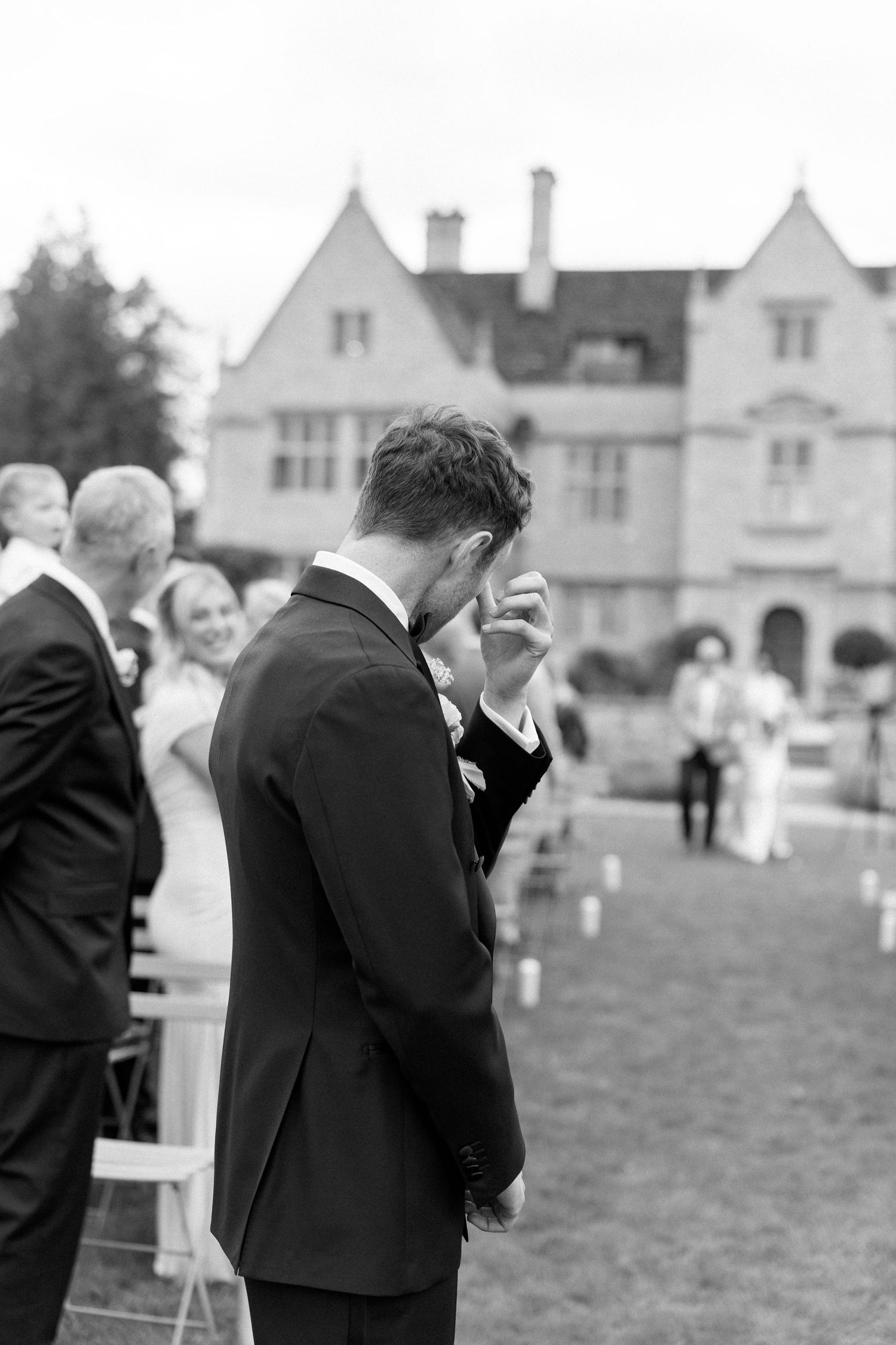Emotional groom wipes tears as bride walks down outdoor aisle at elegant manor wedding.