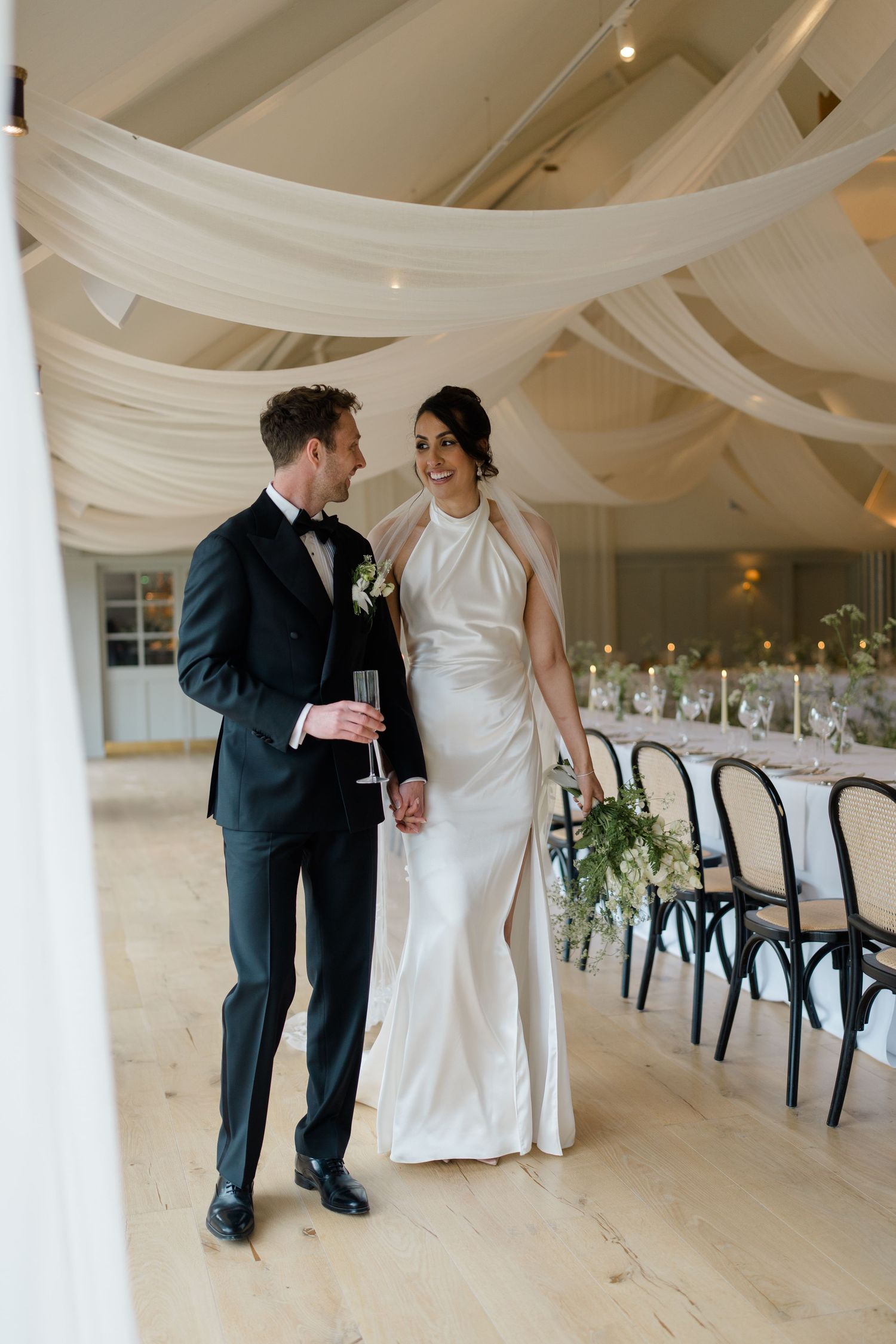 Bride and groom walk through elegantly draped reception venue with long dining table and candlelight.