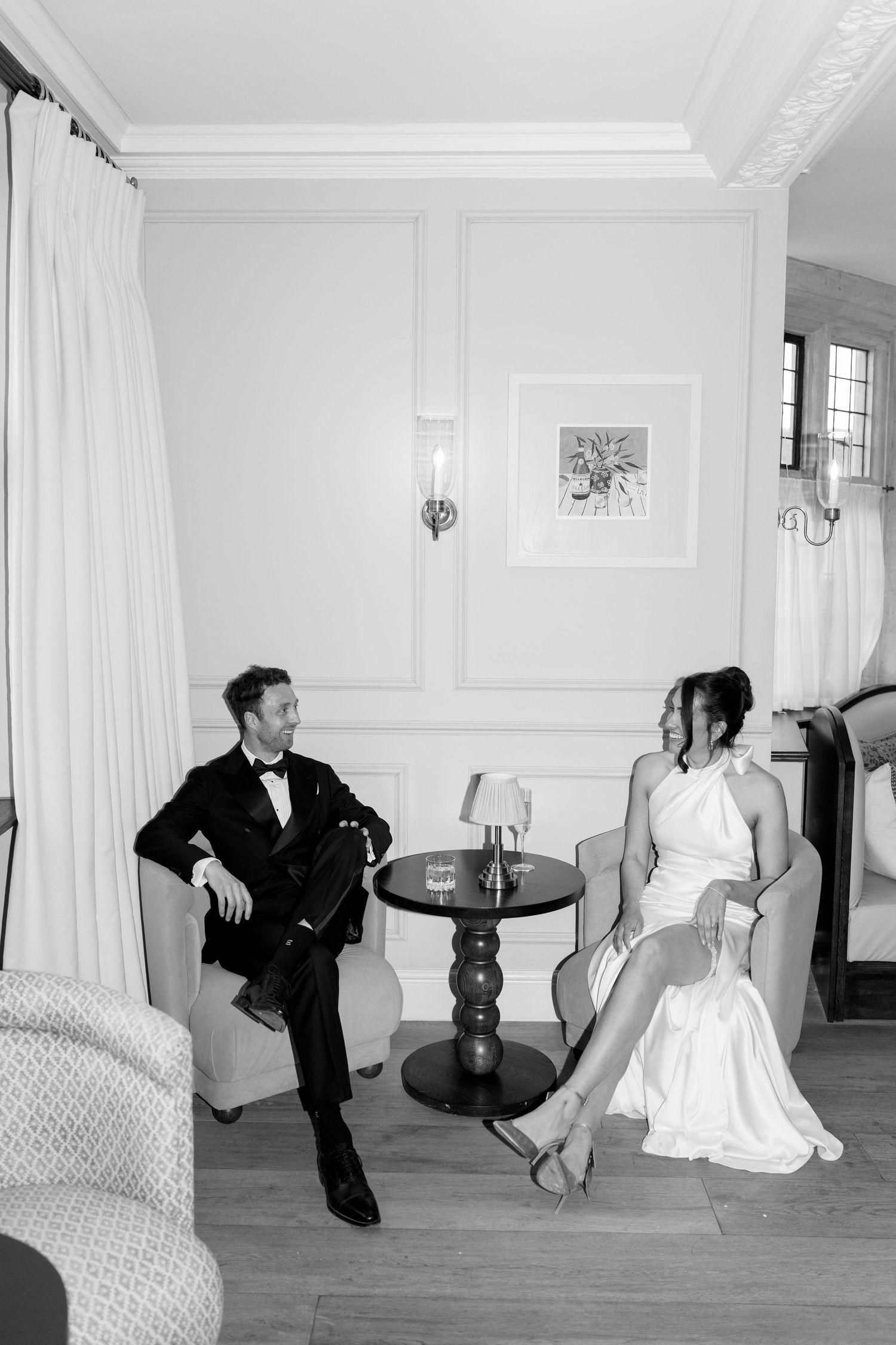 Black and white photo of a bride and groom relaxing in elegant armchairs in a luxury hotel room.