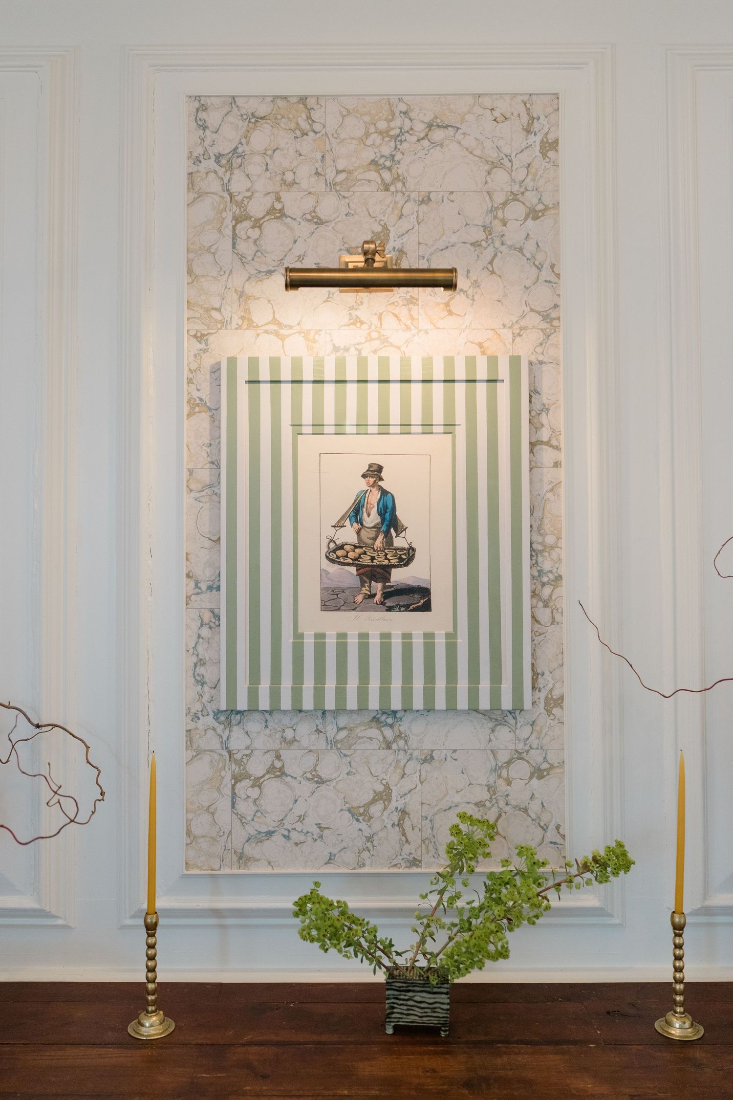 Elegant wall display with striped frame, vintage figure print, marble wallpaper, brass candlesticks, and small green plant on wood console.