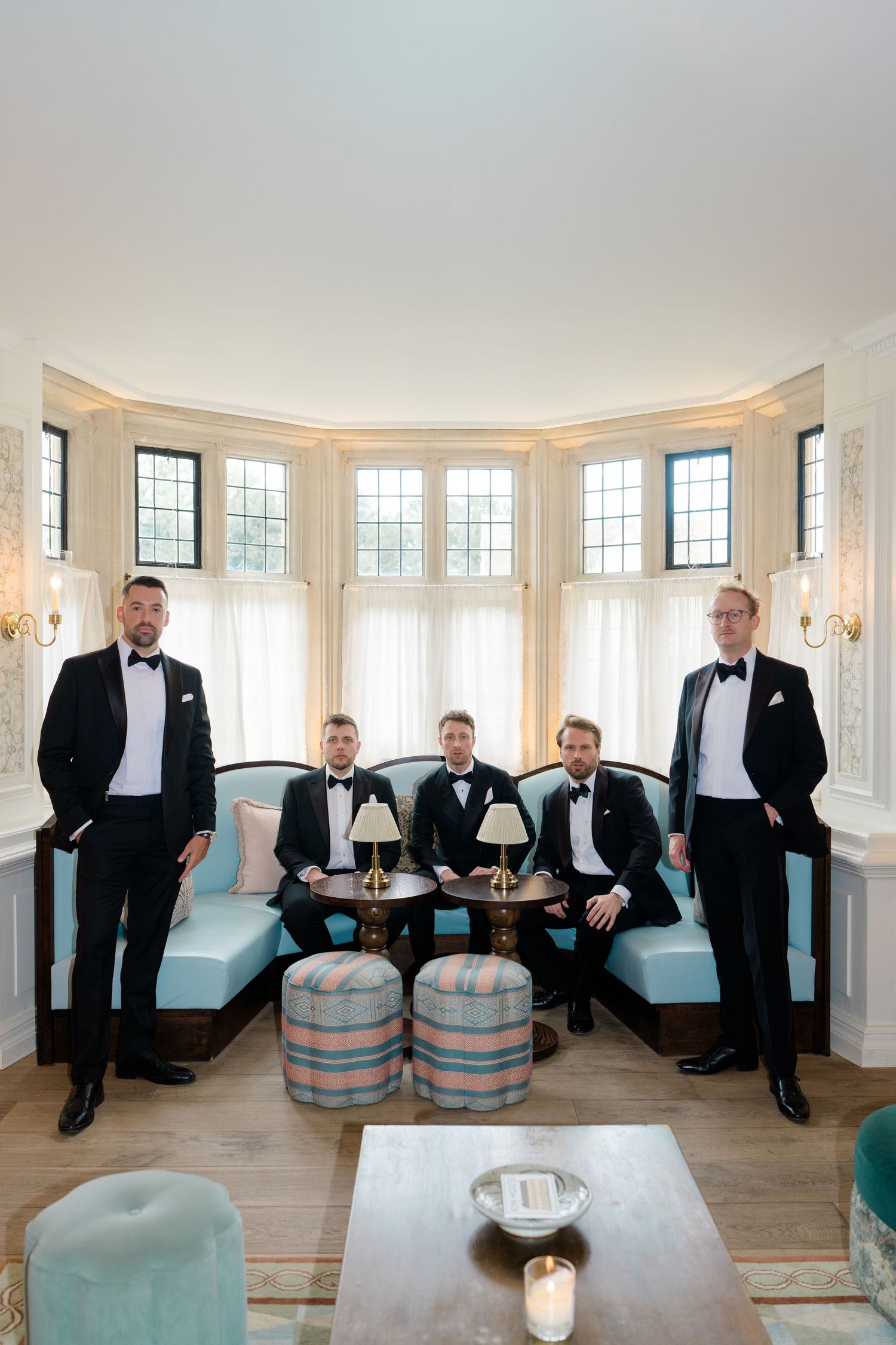 Five groomsmen in black tuxedos pose elegantly in a curved bay window sitting room at a wedding venue.