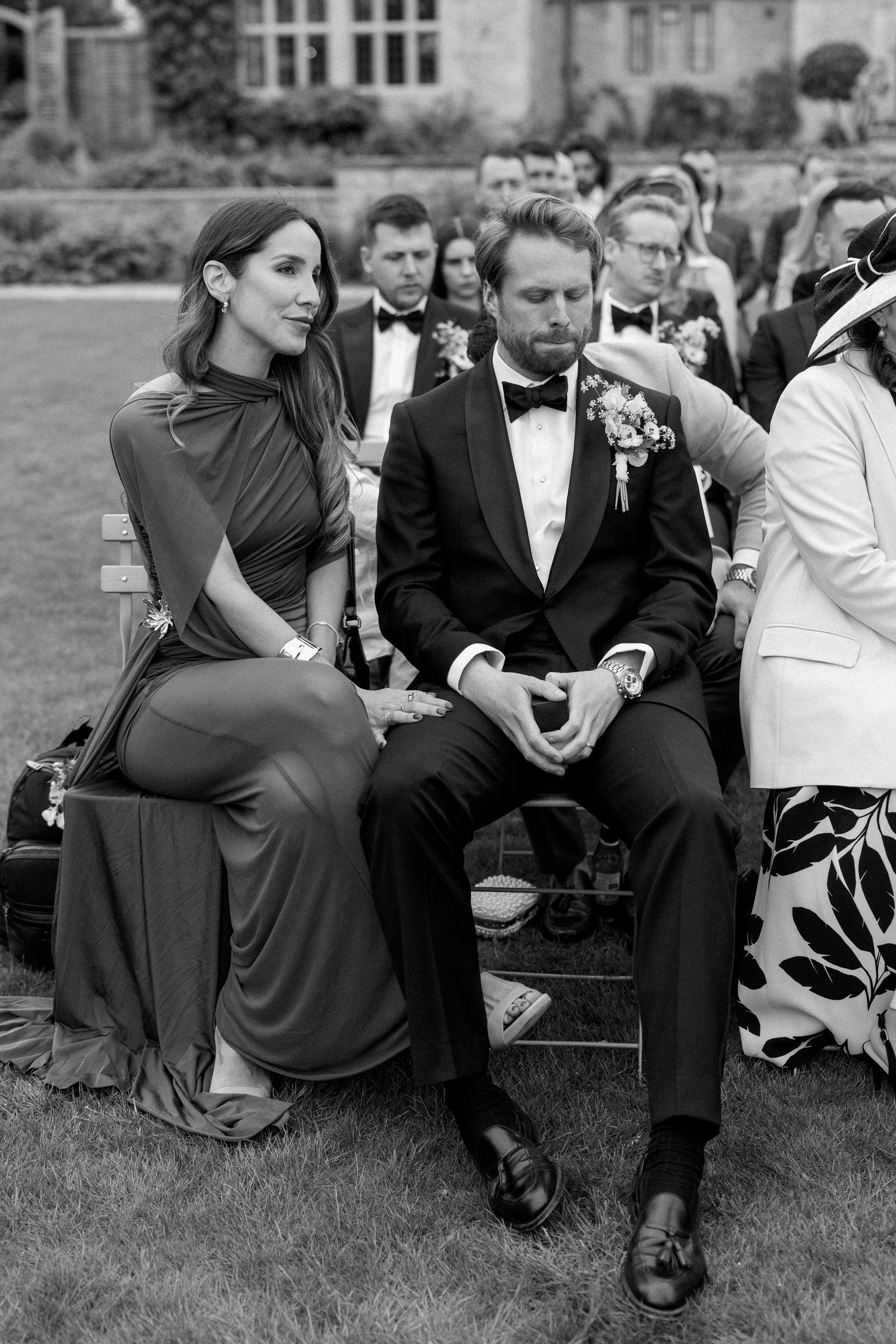 Guests seated at outdoor wedding ceremony, man in tuxedo with boutonniere and woman in elegant gown.