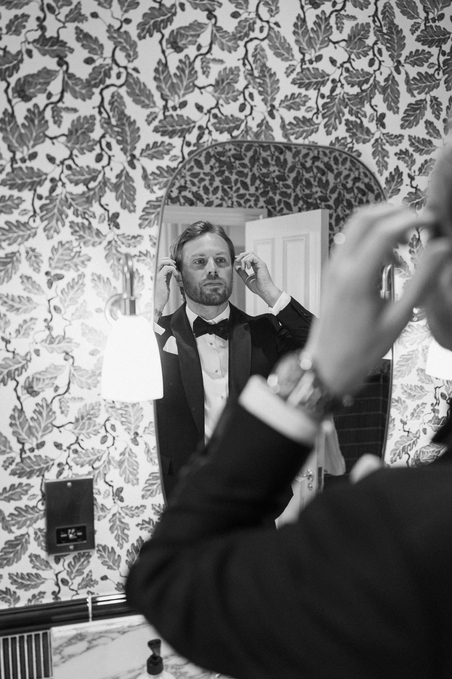 A groom in a tuxedo adjusts his bow tie while looking in a mirror with floral wallpaper backdrop.