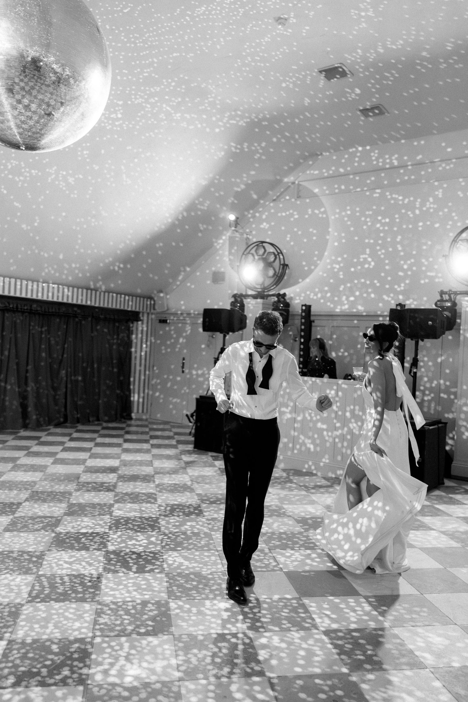 Black and white photo of bride and groom dancing under disco ball lights on checkered dance floor.