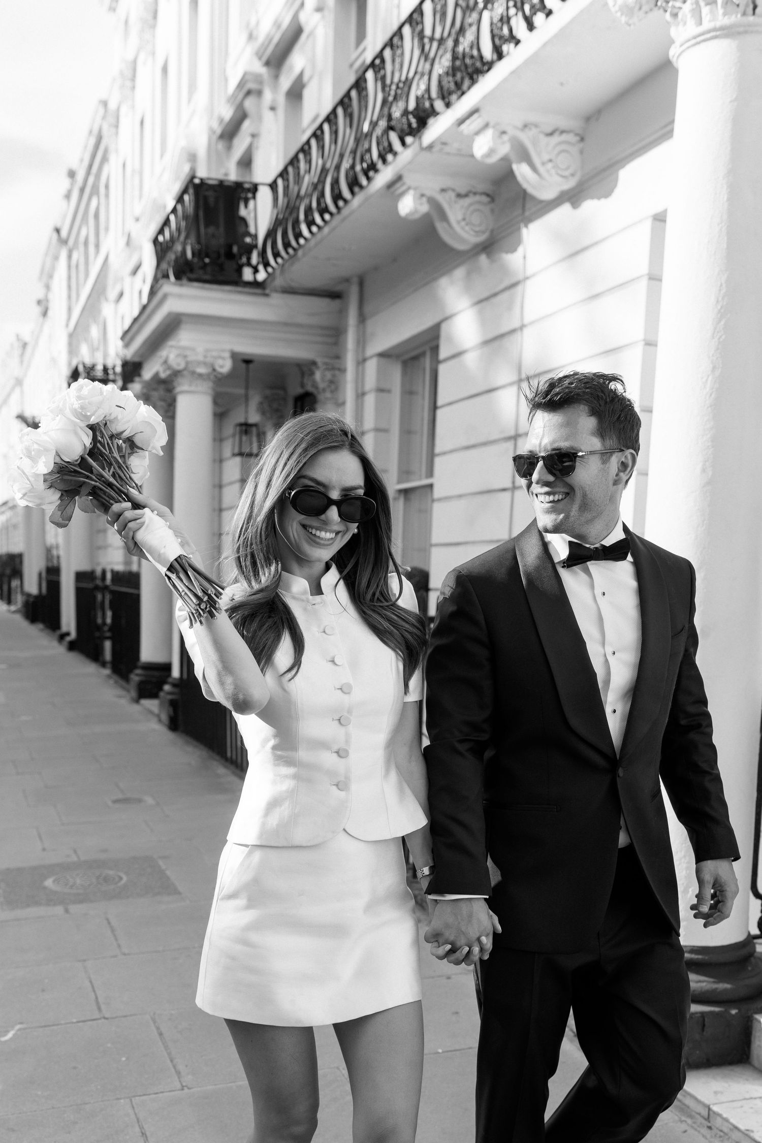 Couple in sunglasses hold hands and bouquet outside a grand London building.