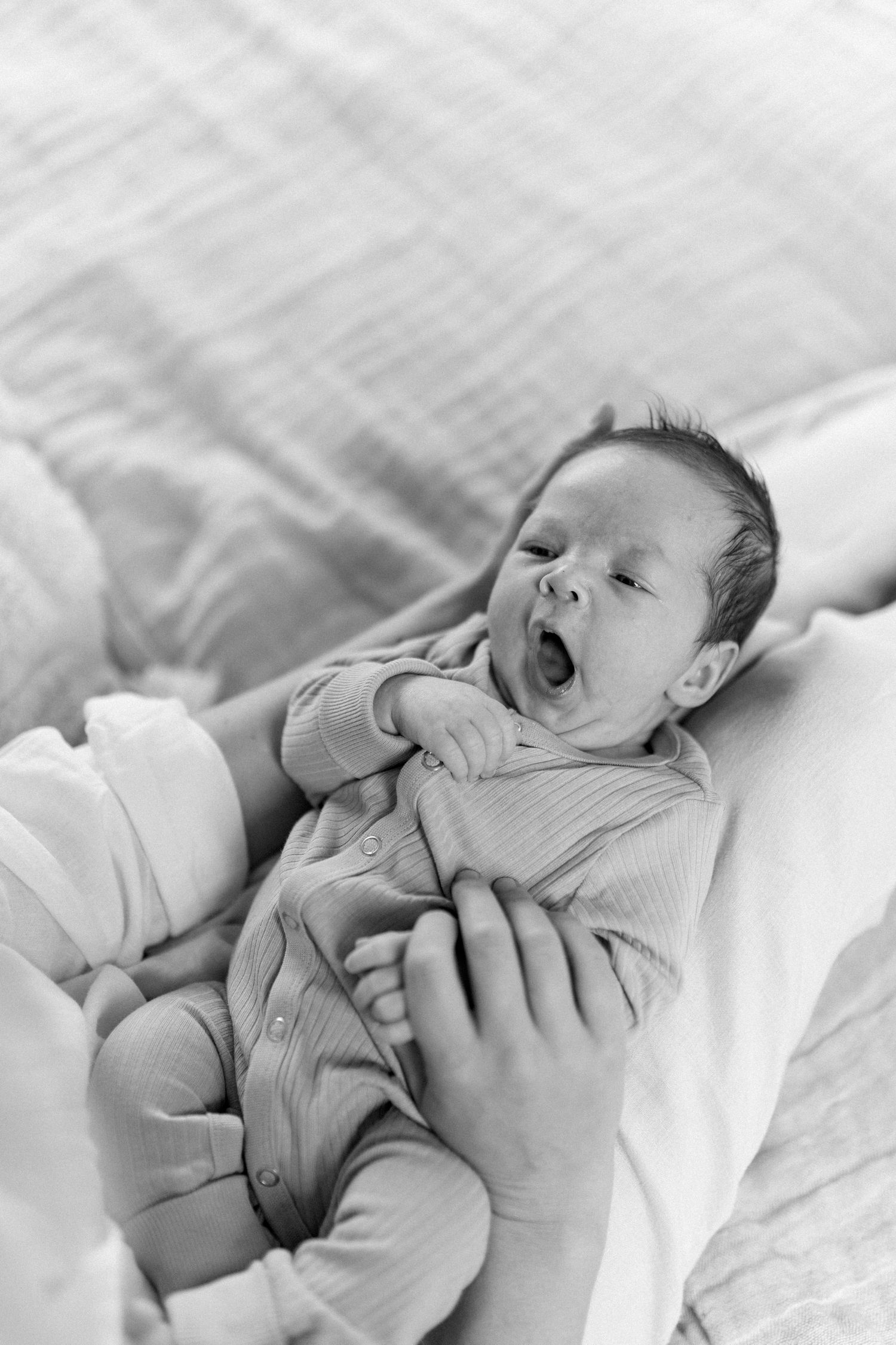 Black and white photo of a sleeping newborn baby yawning while lying on soft white bedding.