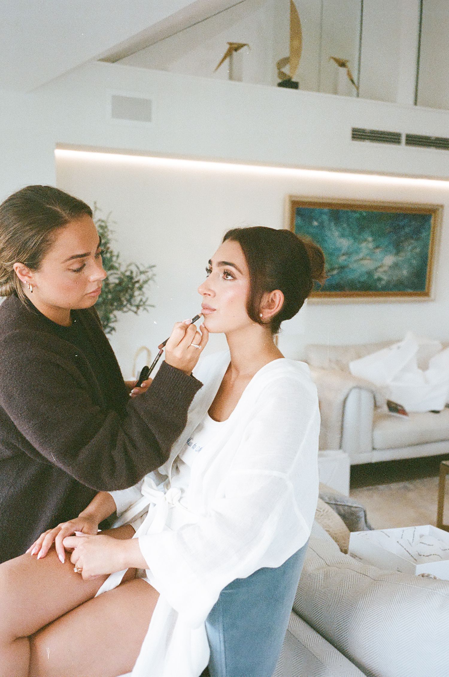 A makeup artist applies cosmetics to a client wearing a white robe in a bright, modern space.