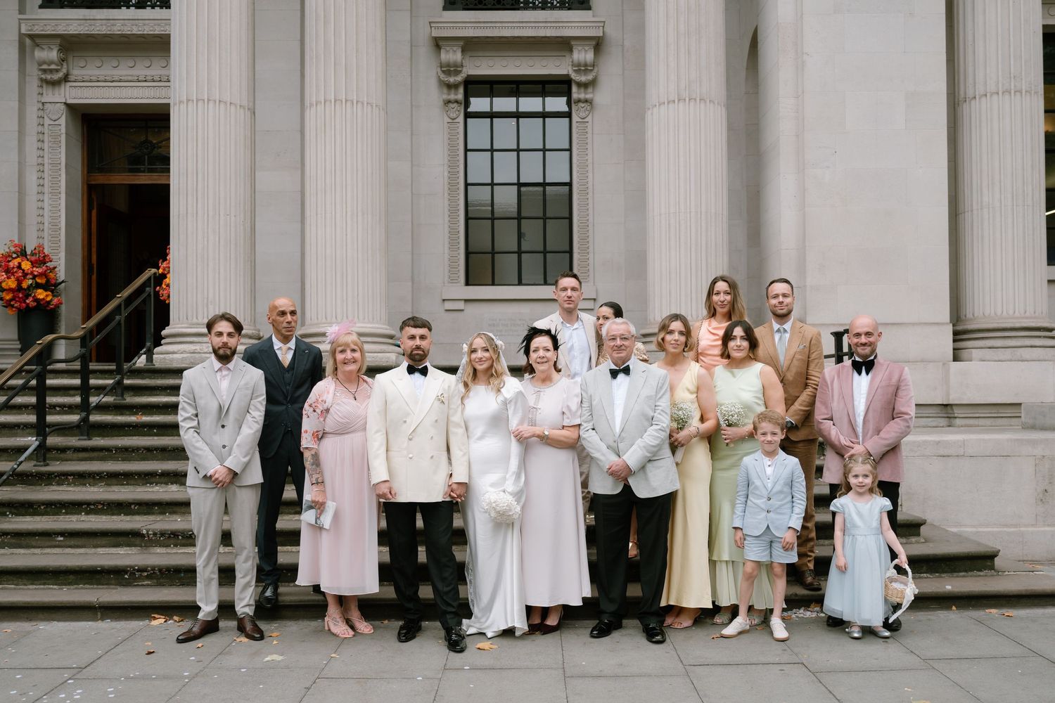 Wedding party poses on grand stone steps of a classical building in coordinated white and pastel attire.