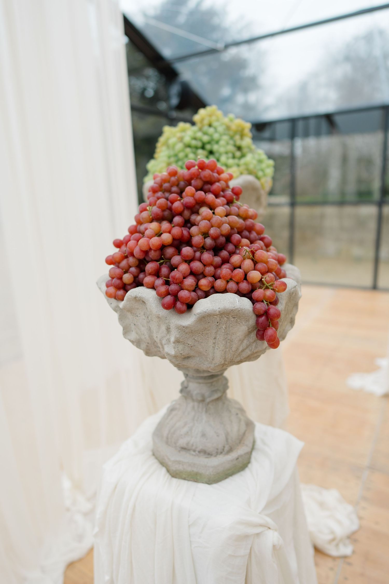 Stone urns filled with red grapes create elegant wedding decor against white fabric backdrop.