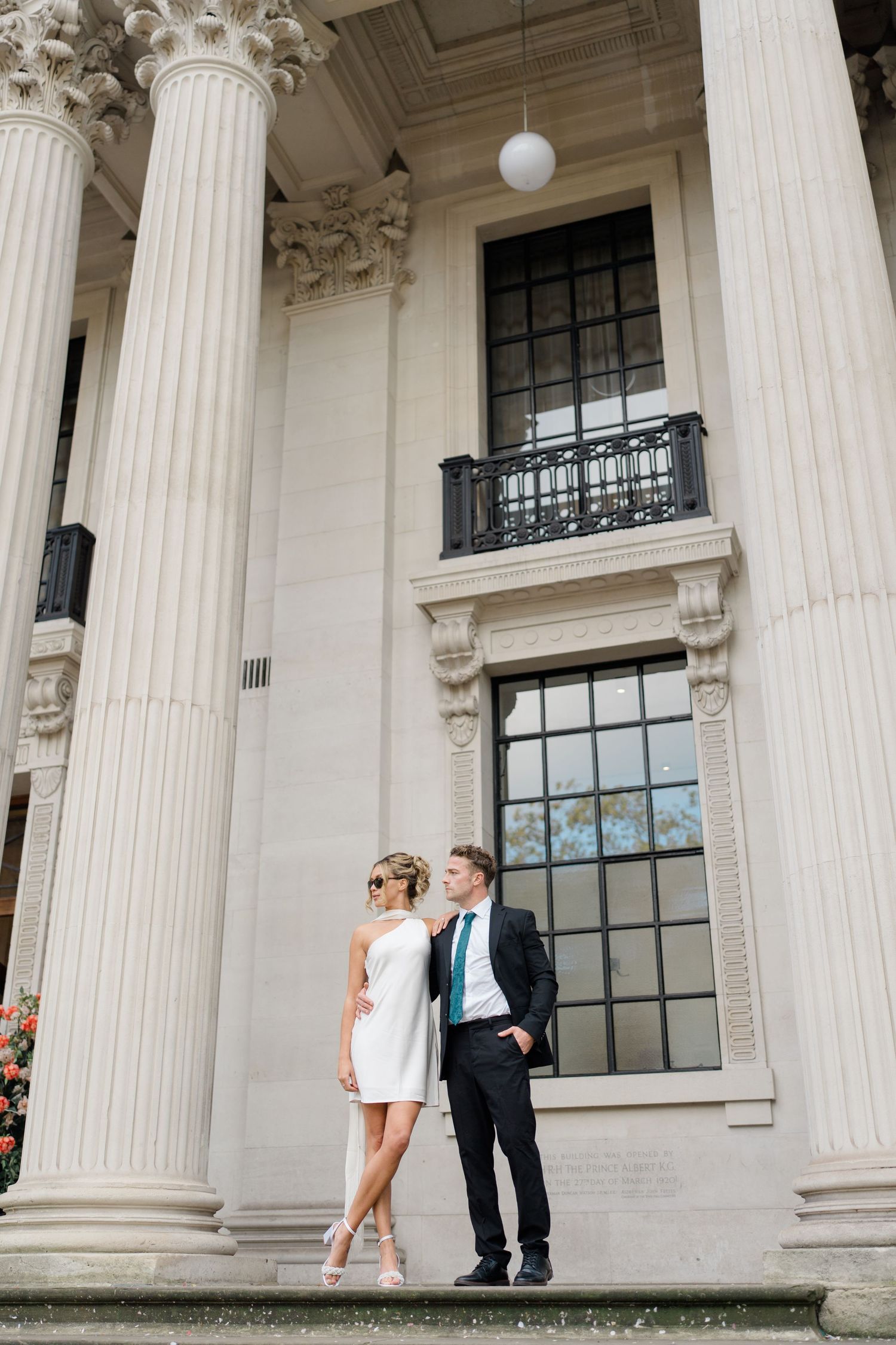 Couple stands between grand white columns of historic building during photo shoot.
