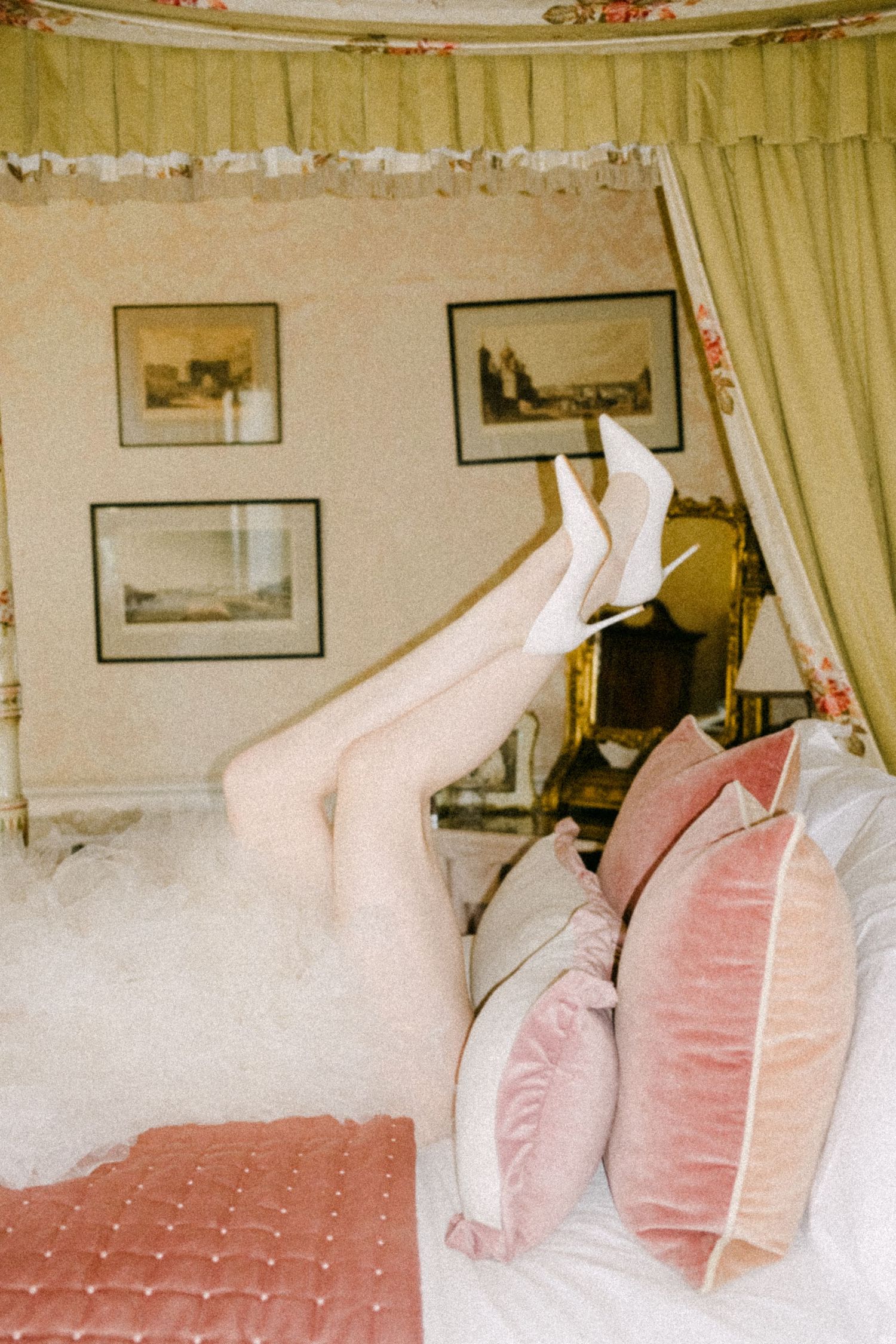 Multiple pillows arranged on a bed in a vintage-style room with framed artwork on cream-colored walls.