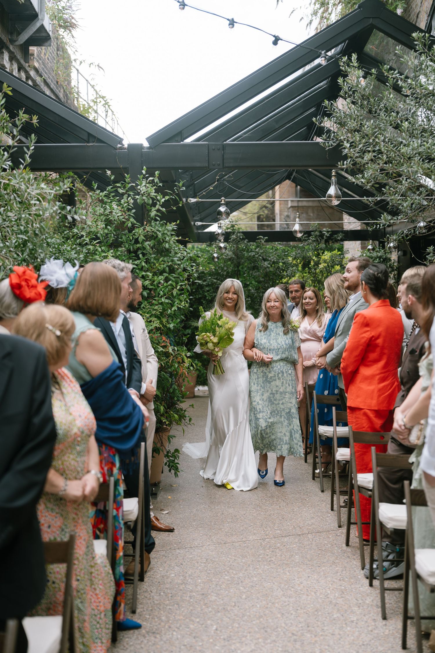 Wedding guests seated in rows under a covered outdoor venue with string lights and greenery.