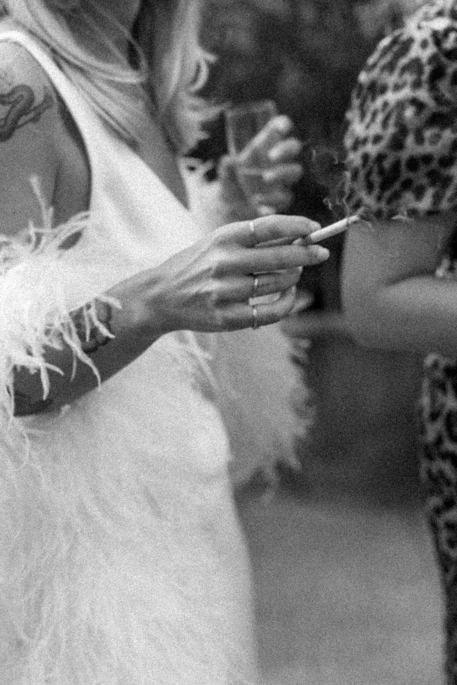 Close-up black and white shot of hands holding a cigarette with feathered dress details visible.