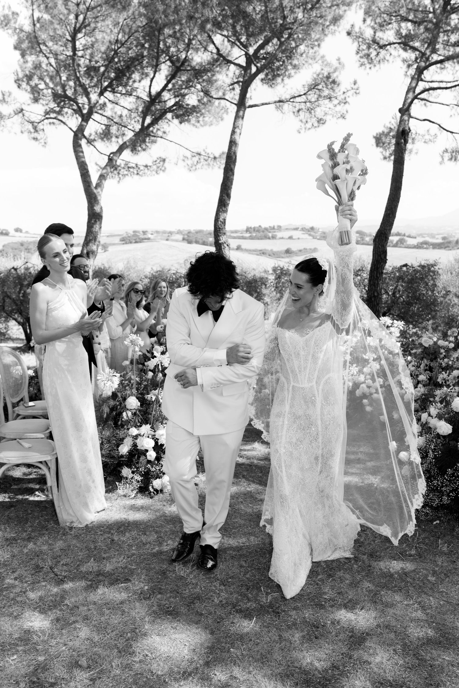 Group dressed in white outfits dancing and celebrating outdoors under pine trees.