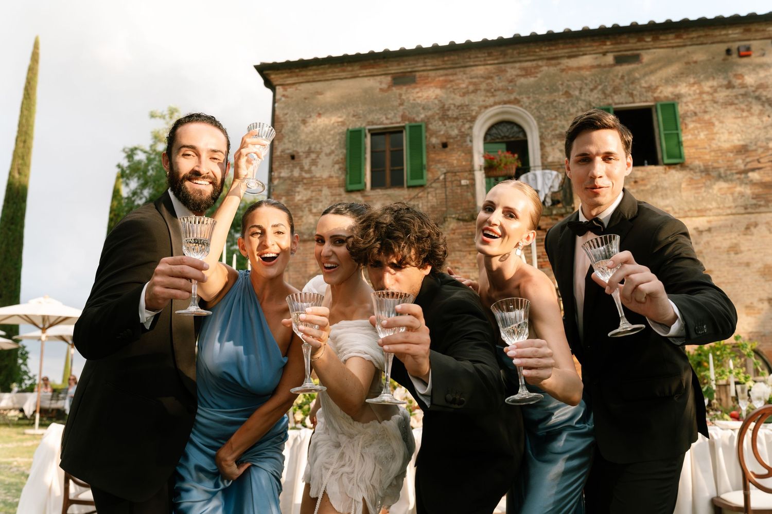 Wedding guests raising glasses in a toast outside a rustic stone building.