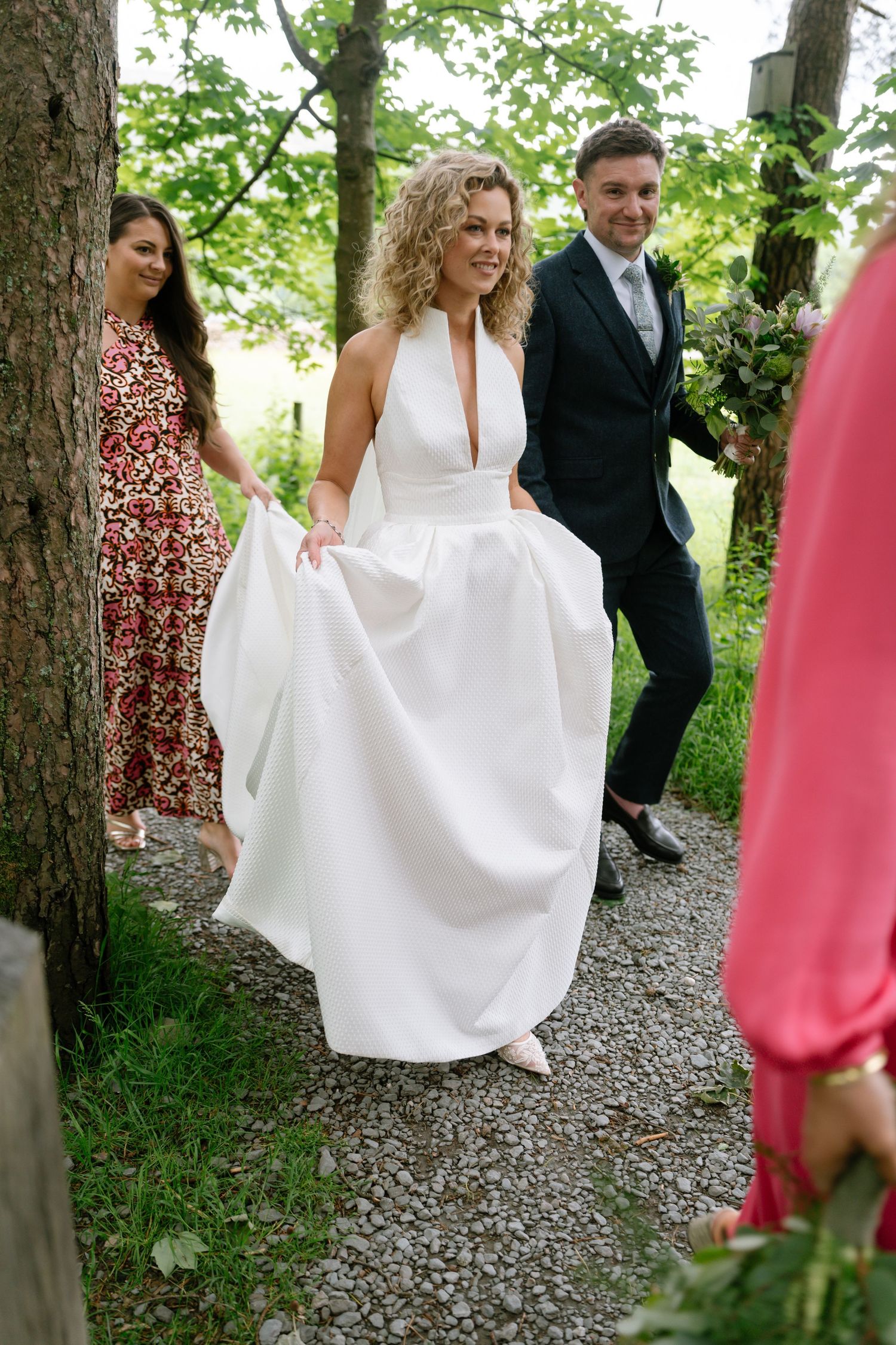 A wedding party walks down a wooded path with the bride in an elegant white halter gown.