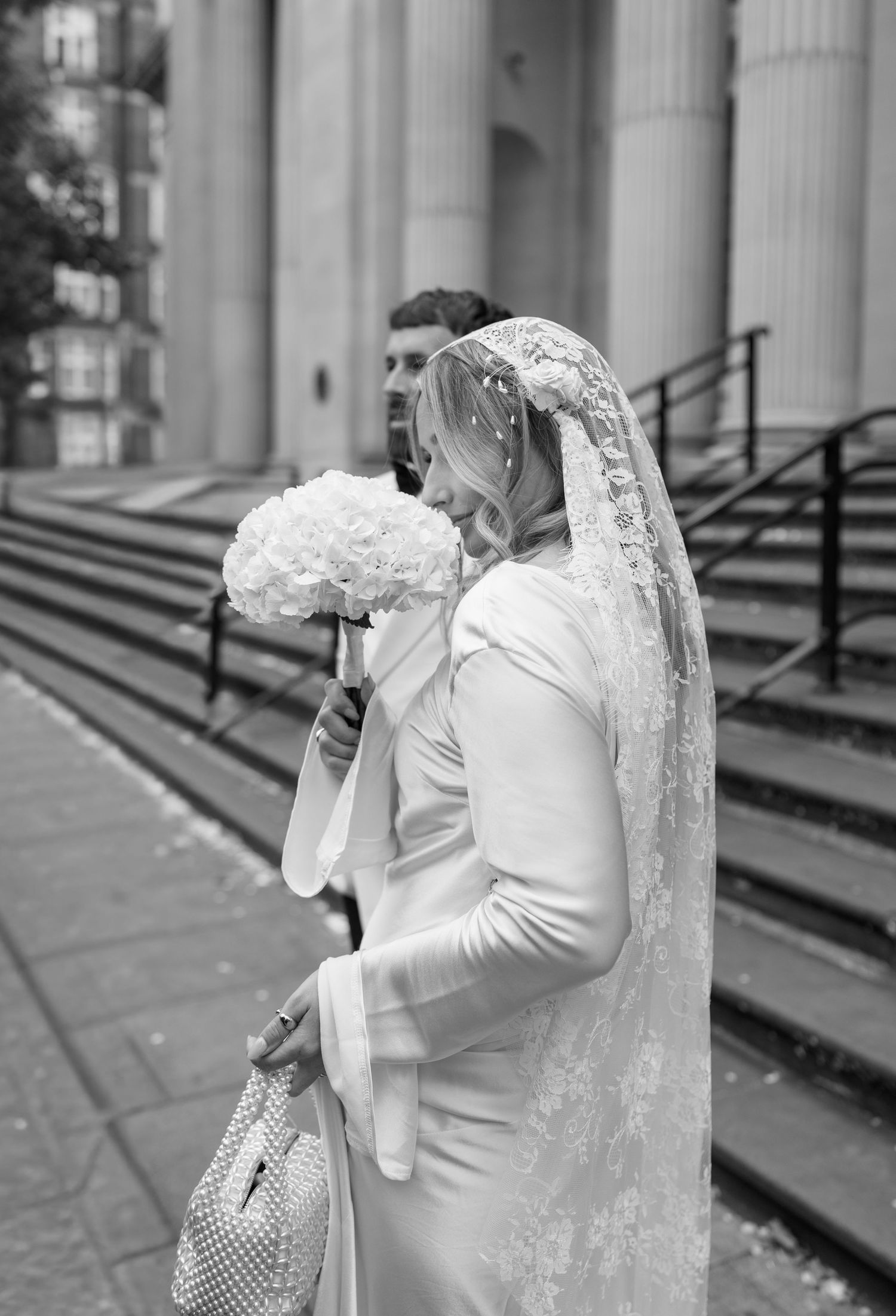 A bride in a long veil holds a white bouquet on church steps.